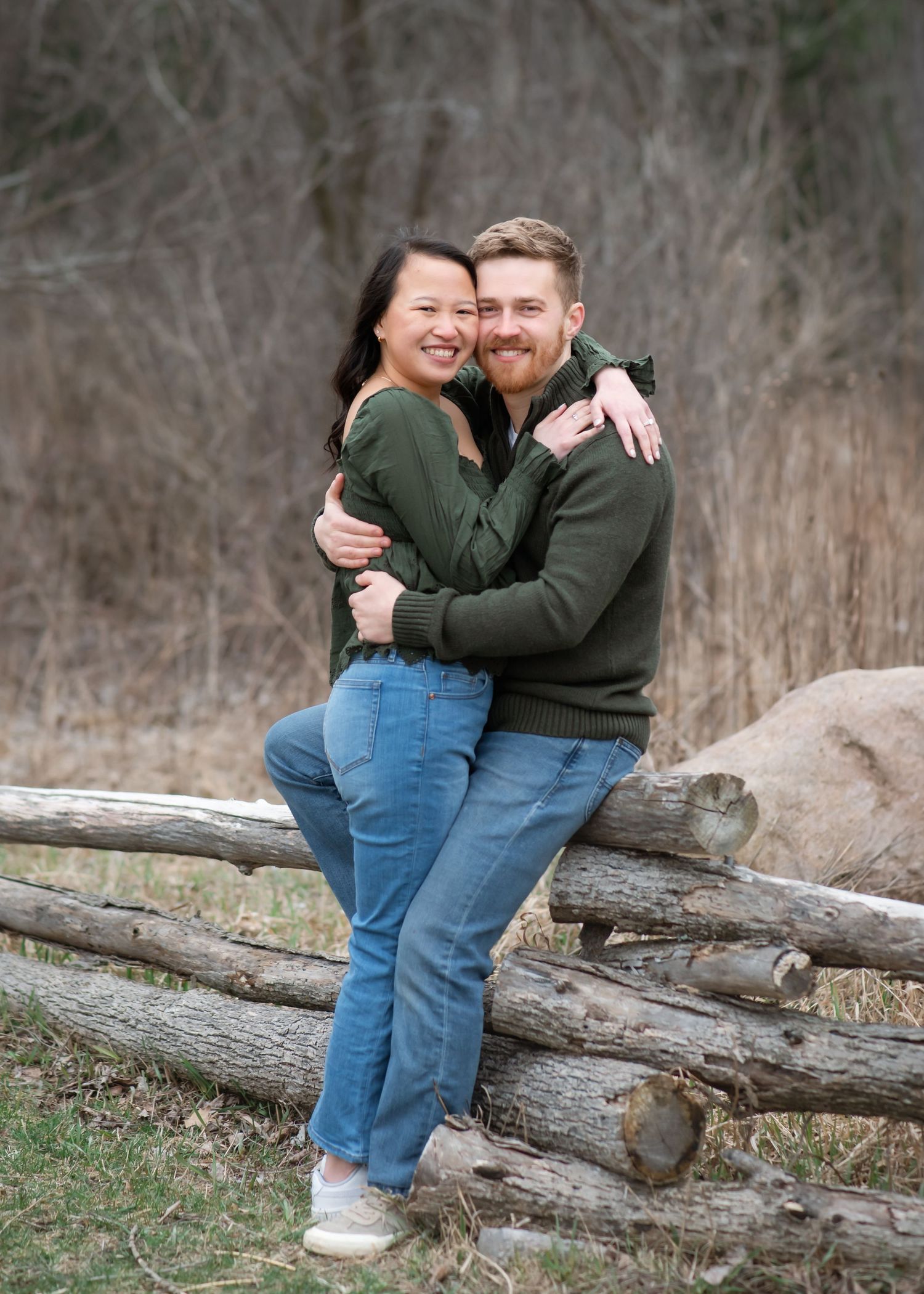 A couple in casual attire shares a tender kiss against a tree trunk in a natural outdoor setting.