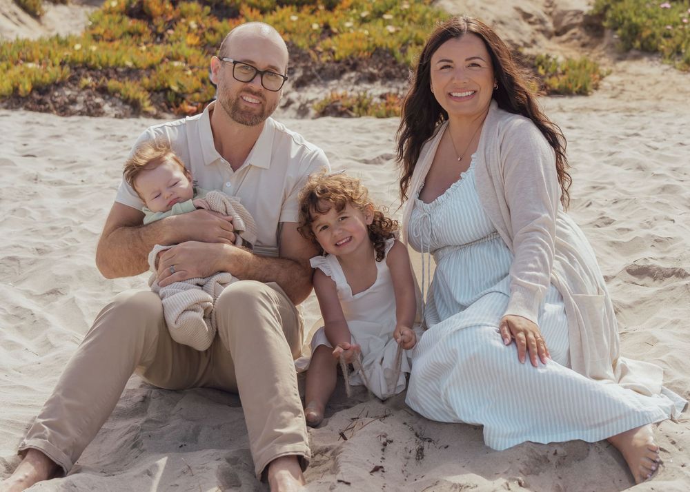A group in neutral colored clothing sits together on a sandy beach during a sunny day.