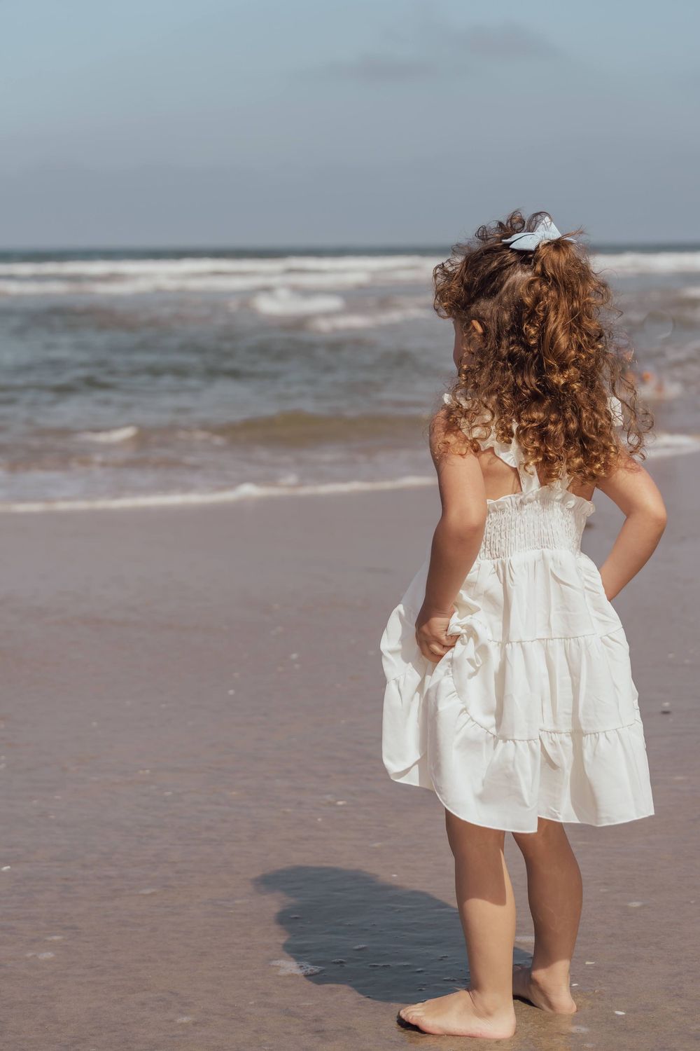 A toddler in a white dress plays at the edge of the ocean waves on a sandy beach.