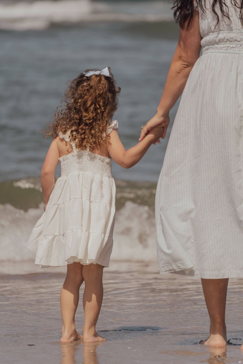 Two people standing barefoot in shallow ocean waves, wearing white clothing at the beach.