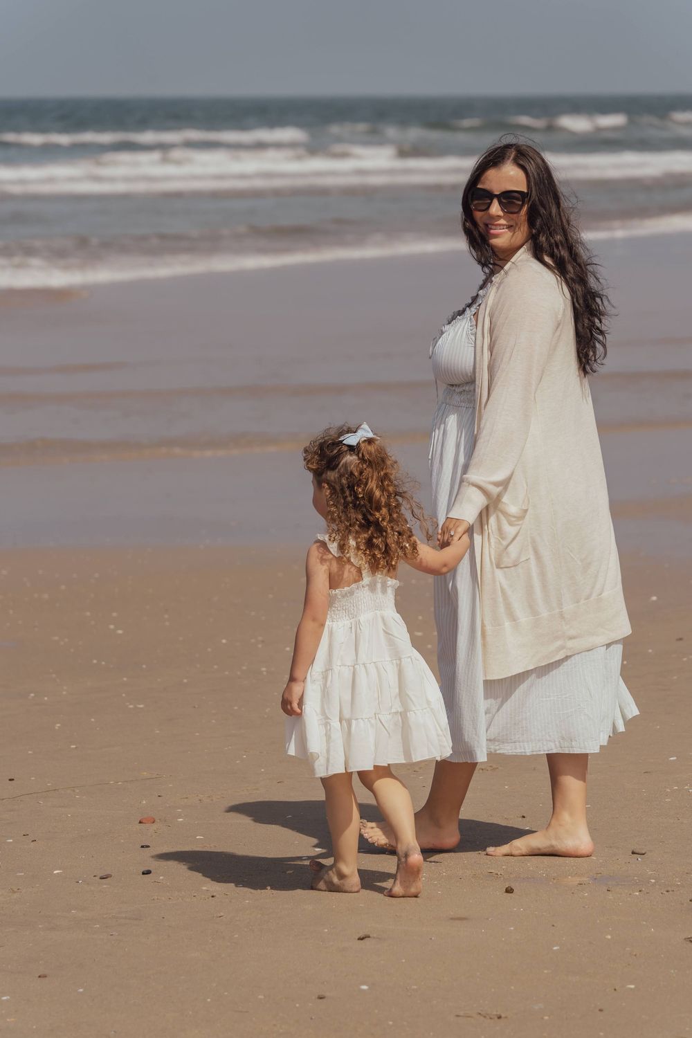 Two people in white dresses walk together along a peaceful beach shoreline.