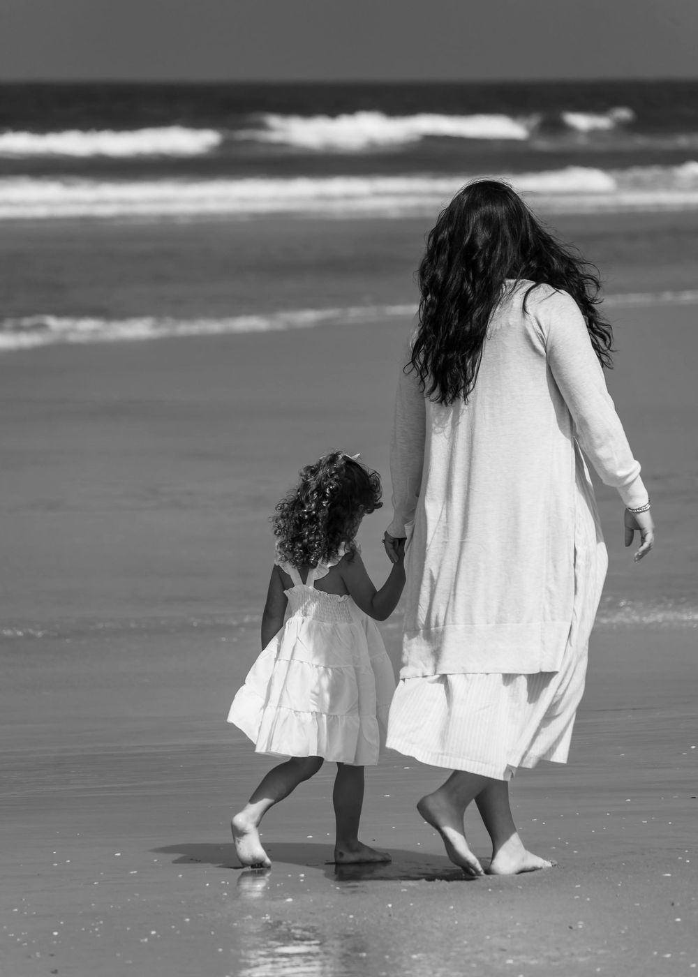 A mother and daughter in white dresses walk hand in hand along a beach shore in black and white.