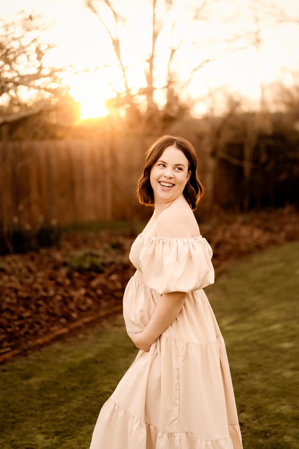 Smiling pregnant woman in cream off-shoulder dress poses outdoors at golden sunset in a lush backyard garden.
