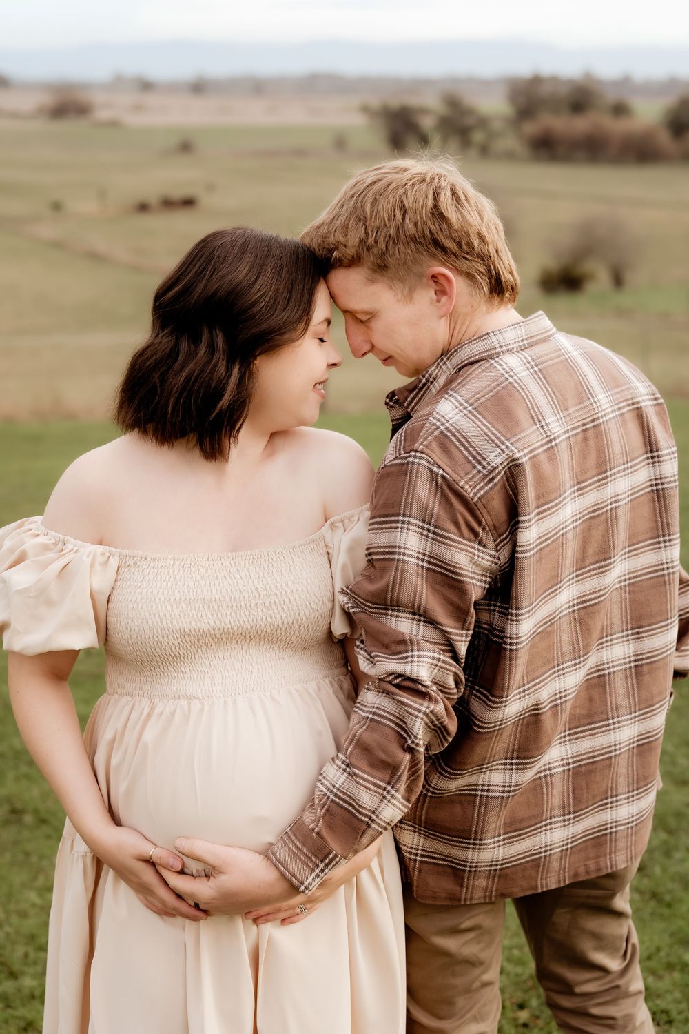 A couple shares a tender moment outdoors, the pregnant woman in a cream dress cradling her bump as her partner leans in close.