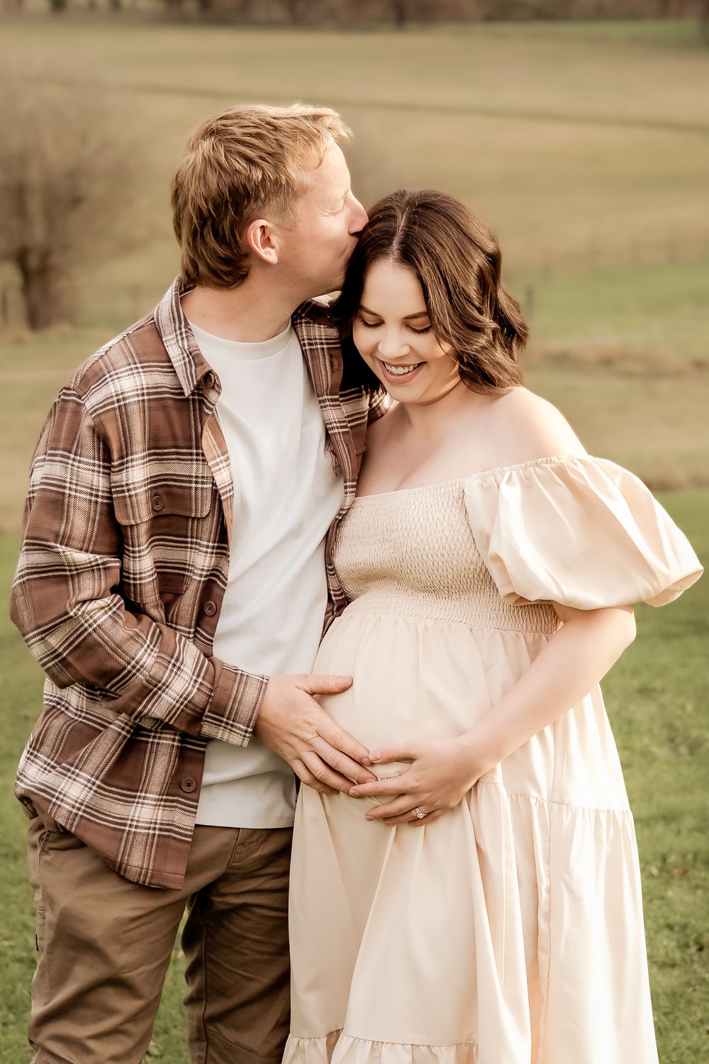 A man kisses a smiling pregnant woman in a cream off-shoulder dress outdoors on a green field.