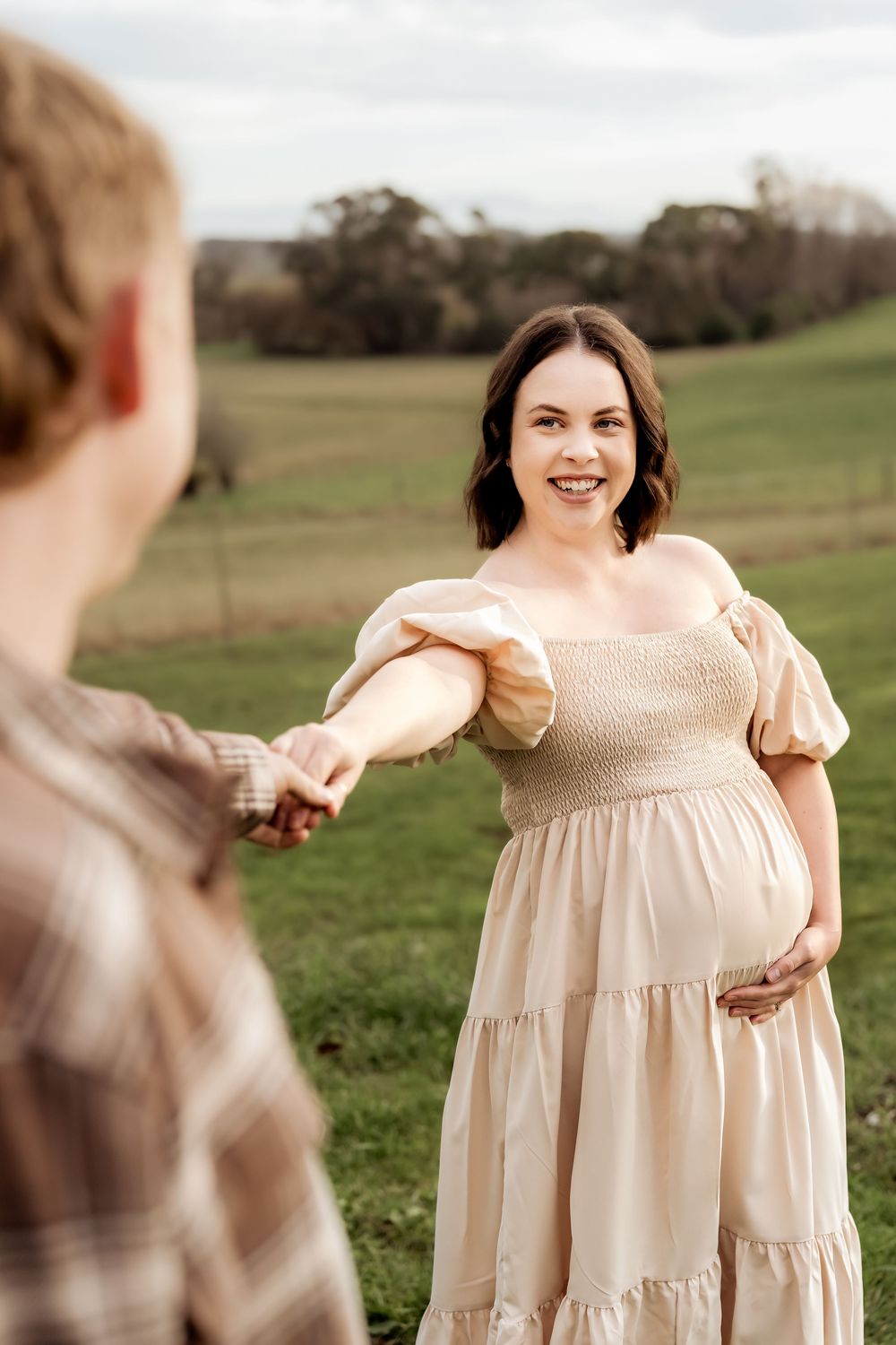 Smiling pregnant woman in beige dress holds partner's hand in green field during outdoor maternity photoshoot.