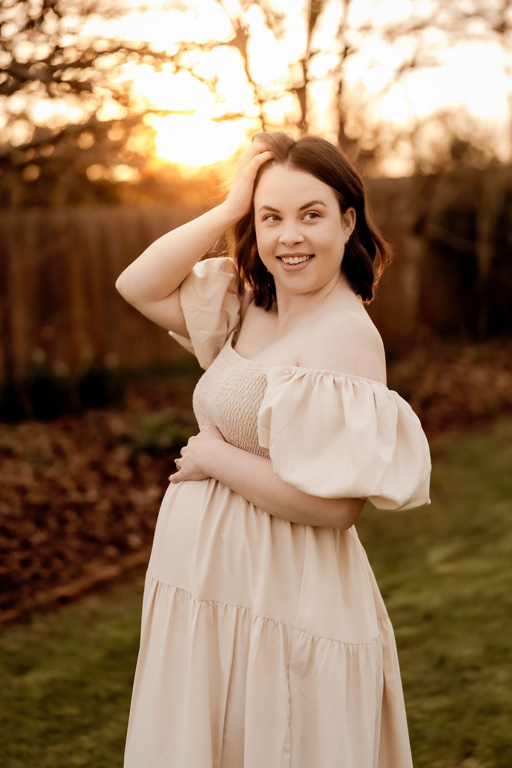 Smiling pregnant woman in cream off-shoulder puff sleeve dress poses at golden sunset in outdoor garden setting.