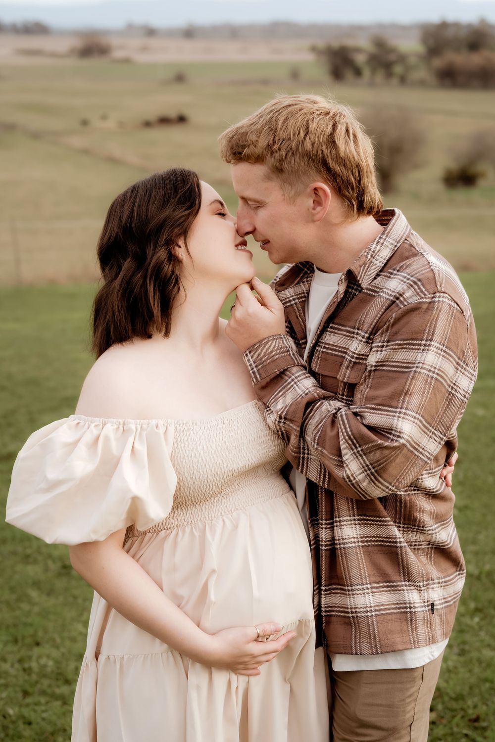 A pregnant couple shares a tender kiss in a lush green field, the woman in a white off-shoulder dress.