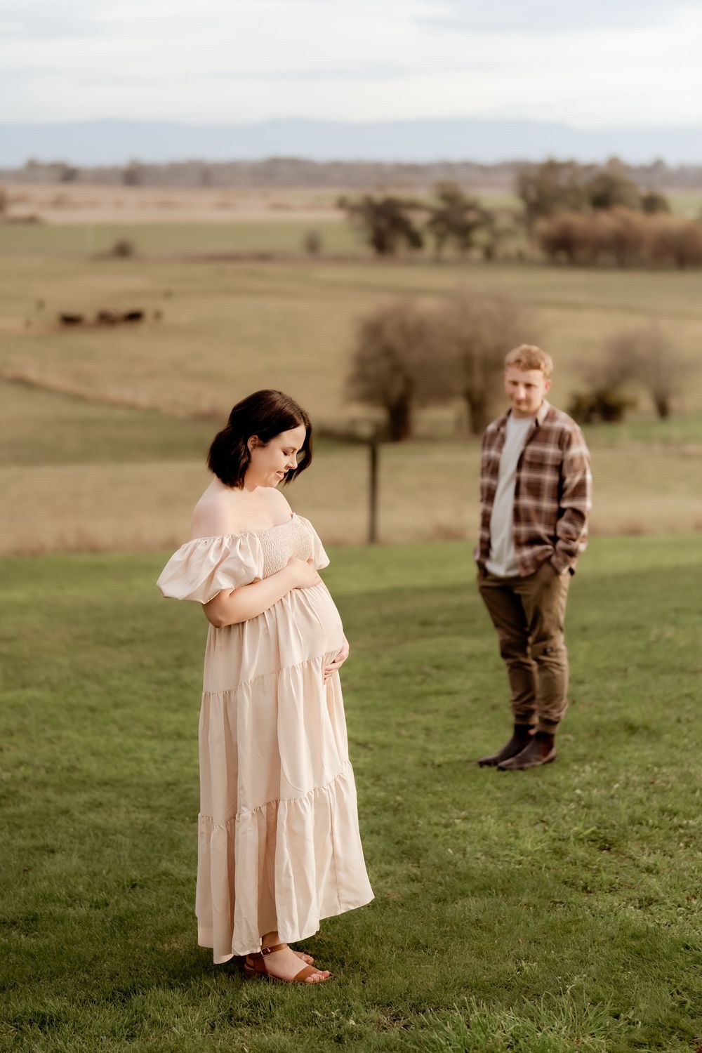 Pregnant woman in cream off-shoulder dress cradles bump in green field, man in plaid shirt stands behind her.