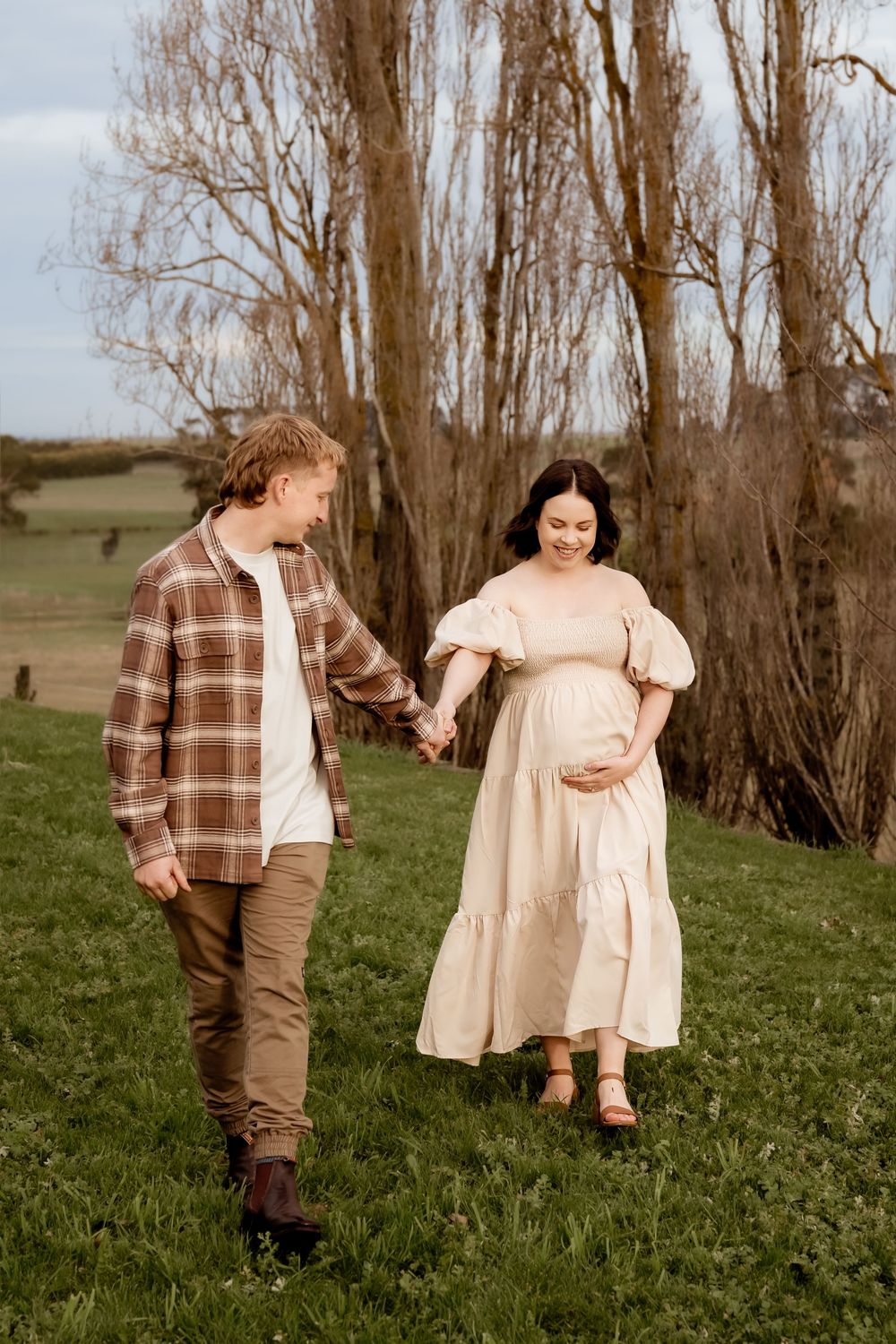 Pregnant woman in cream dress holds hands with man in plaid shirt while walking on green grass near bare trees.