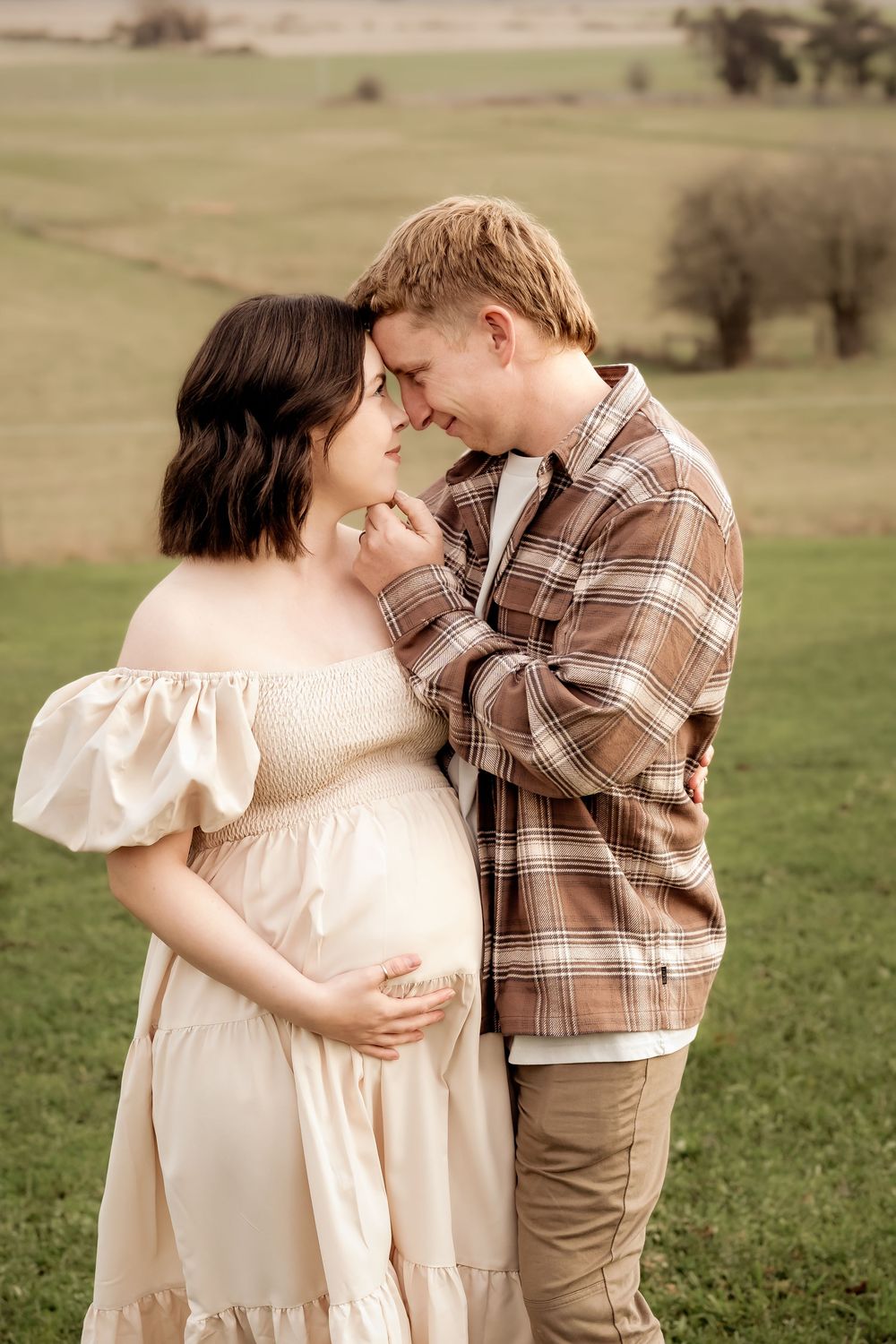 A pregnant couple shares a tender forehead-to-forehead moment outdoors, she in a cream dress, he in a plaid shirt.