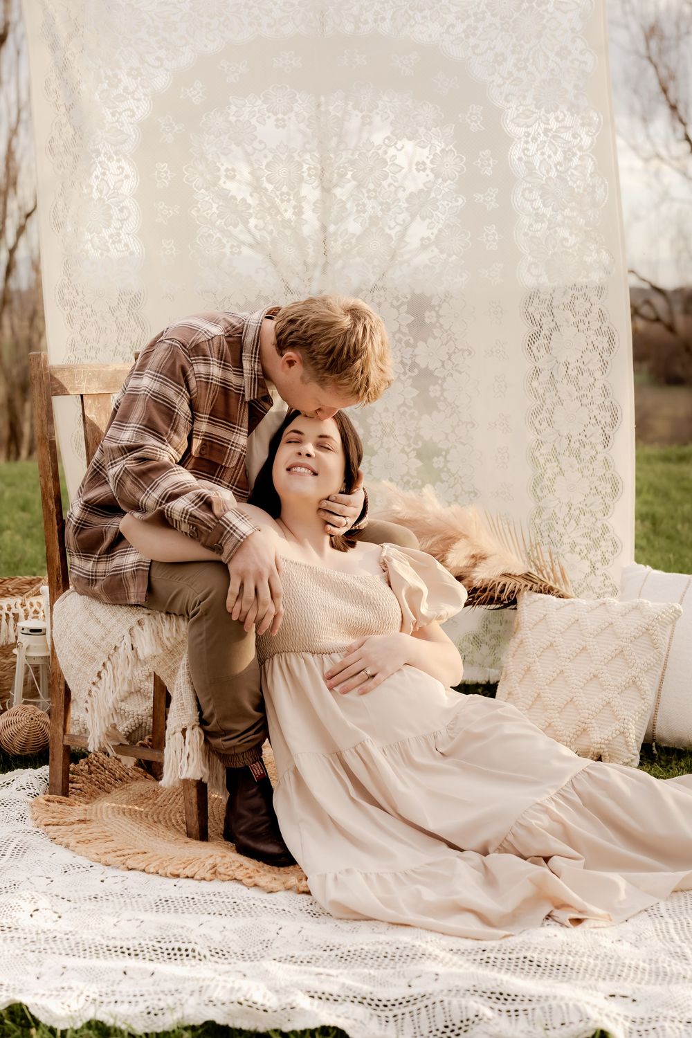 A man kisses a smiling pregnant woman in a boho outdoor maternity photo shoot with lace backdrop.