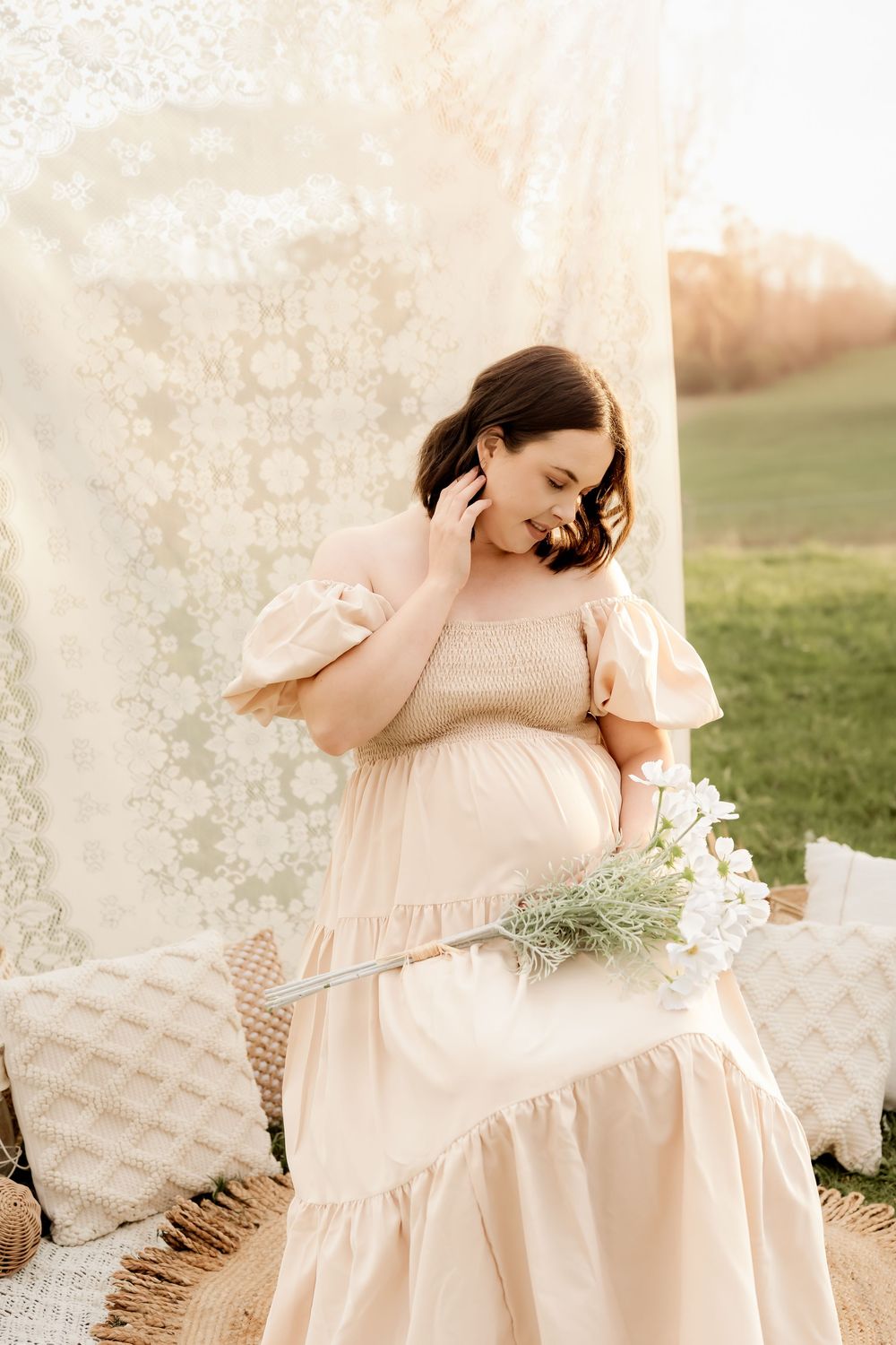 Pregnant woman in peach dress holding white flowers outdoors with lace backdrop in golden sunlight.