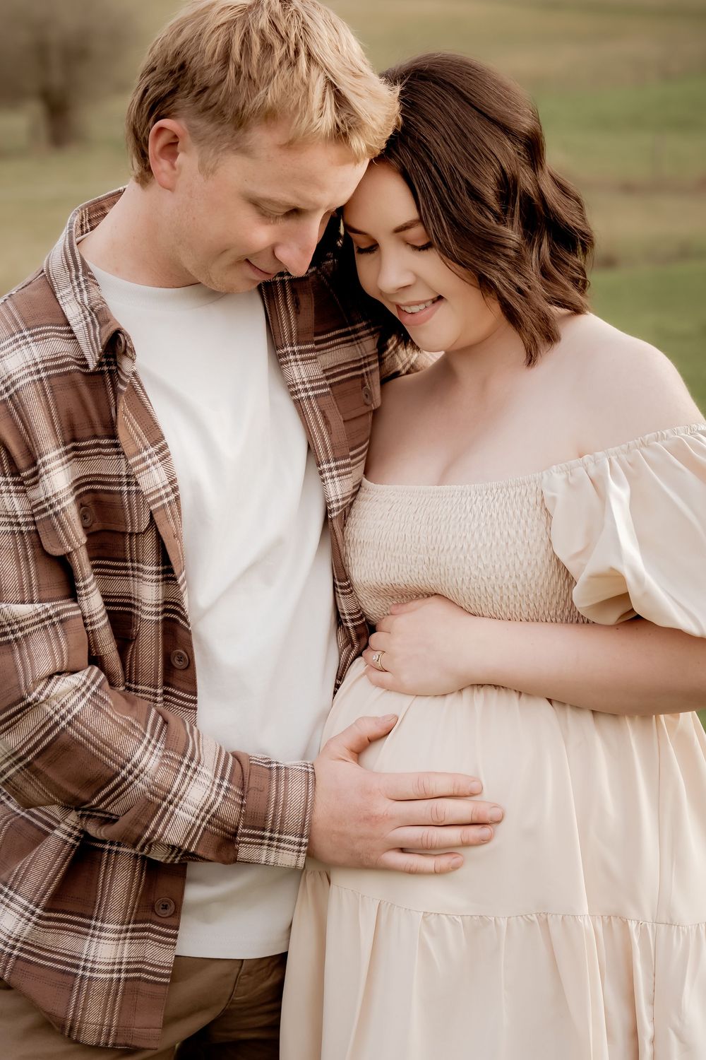 A couple tenderly cradles a pregnant belly outdoors, the woman in a cream dress and man in a plaid shirt, sharing a joyful moment.