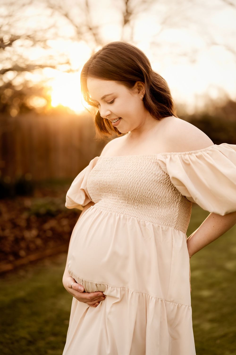Pregnant woman in cream off-shoulder dress cradling baby bump at golden hour sunset outdoors.