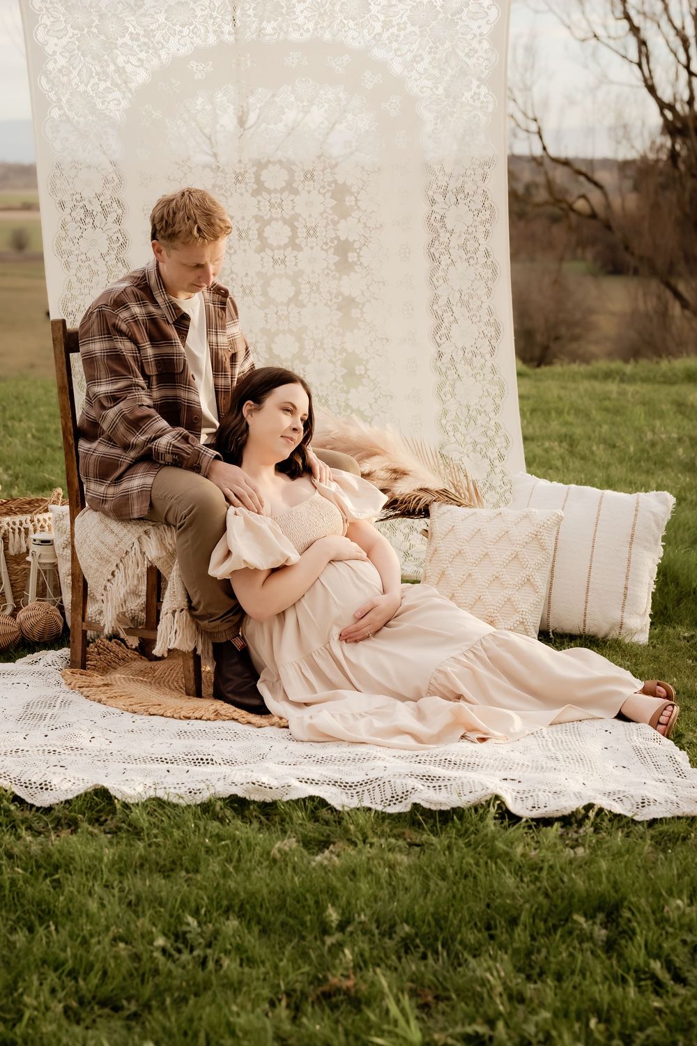 Couple at outdoor boho maternity photoshoot on lace blanket with lace backdrop, pregnant woman in blush dress cradling belly.