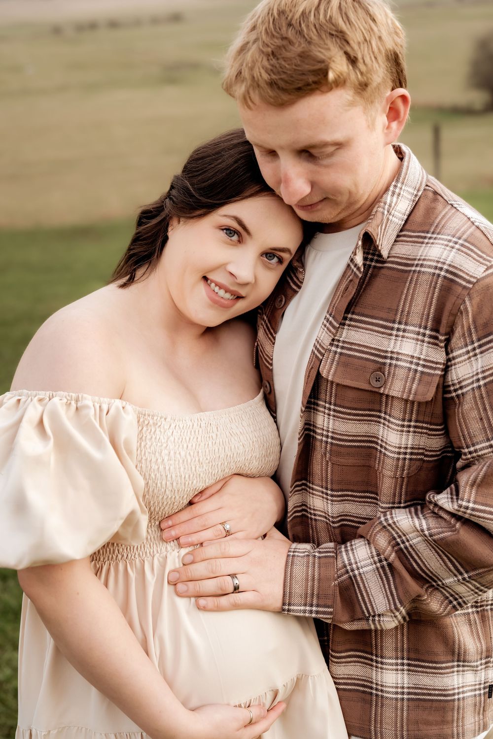 A smiling pregnant woman in a cream off-shoulder dress embraces her partner in a plaid shirt outdoors in a green field.