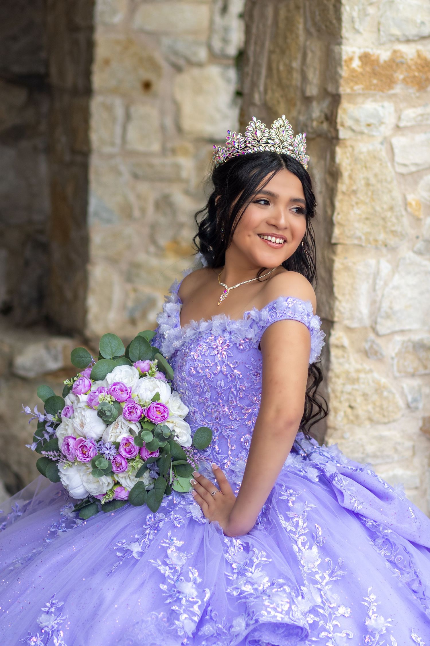 Quinceañera portrait featuring purple ball gown and tiara against stone wall.