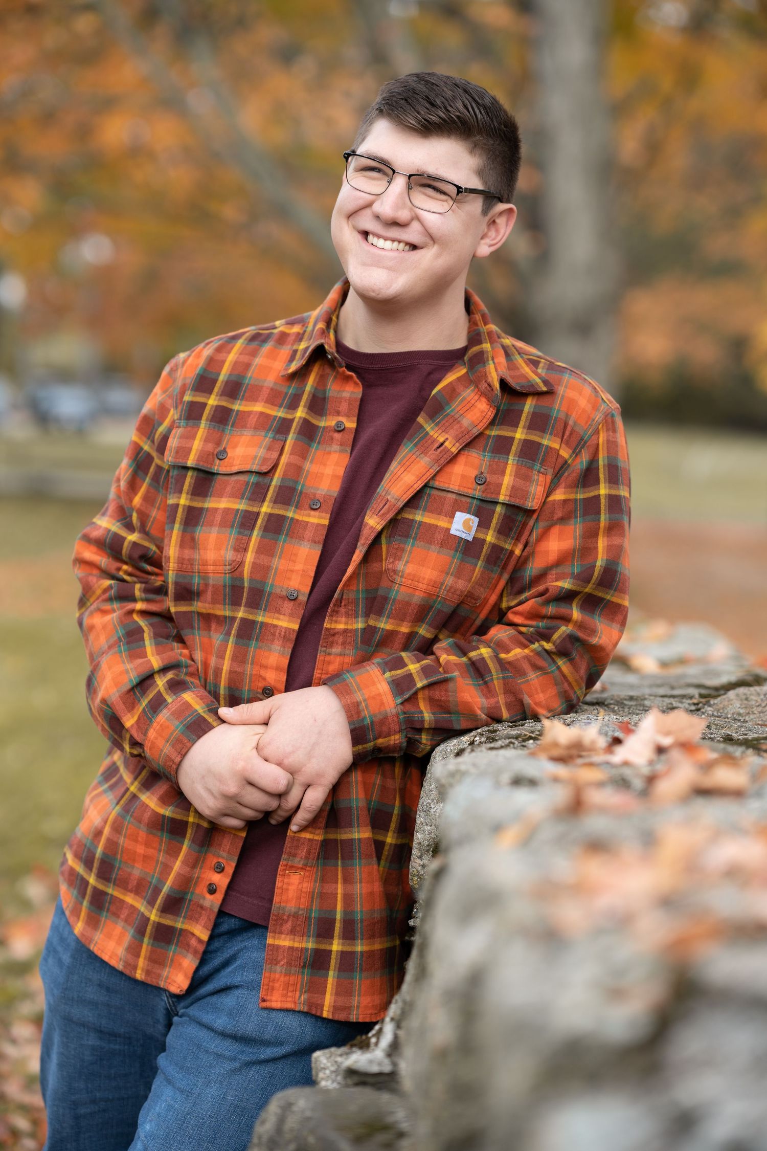 Someone in an orange plaid flannel shirt and glasses poses outdoors against autumn foliage and stone wall.