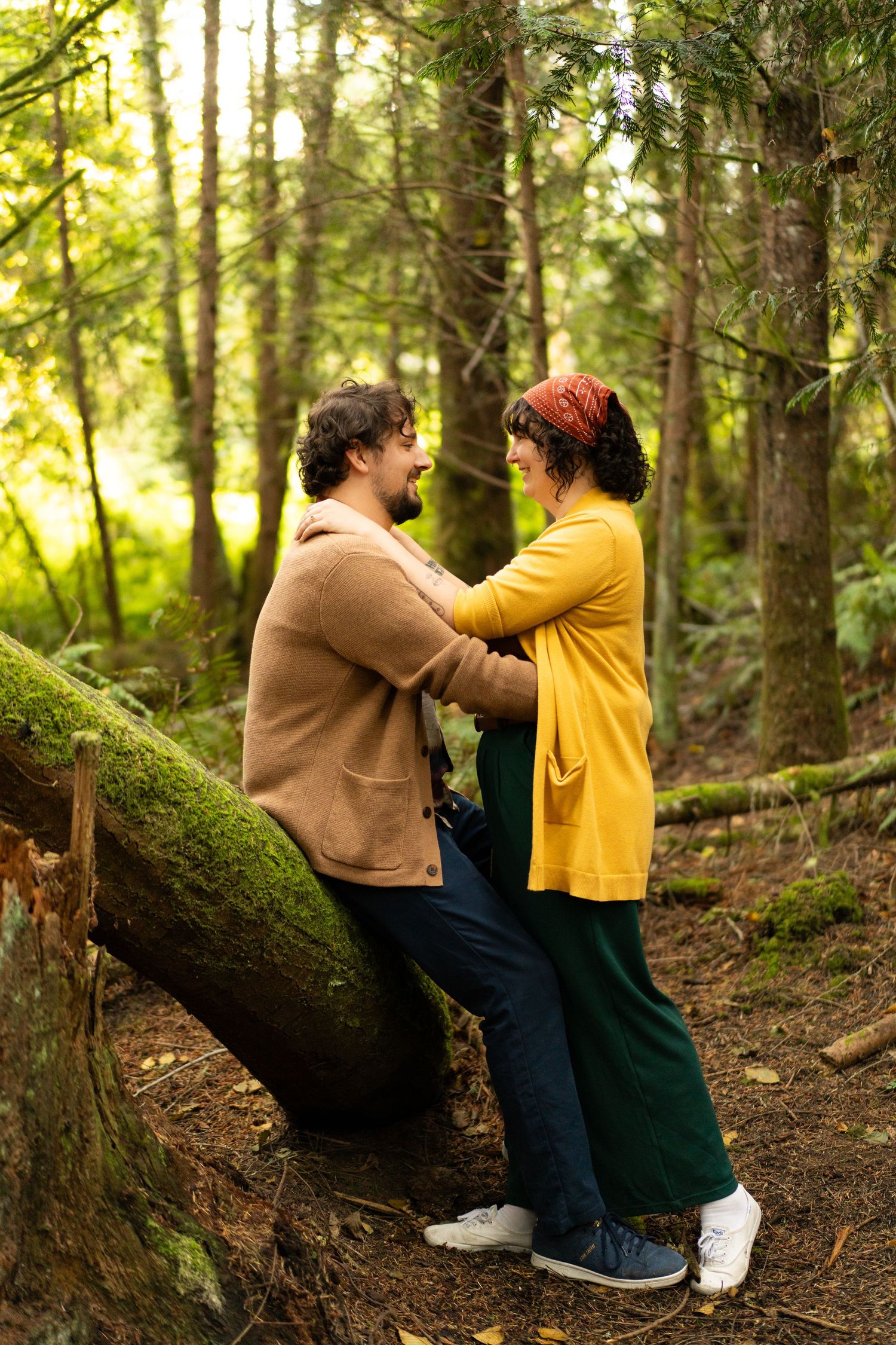 A couple in autumn clothing share an intimate moment while standing on a mossy log in a sunlit forest. Captured by Savvy Photography.