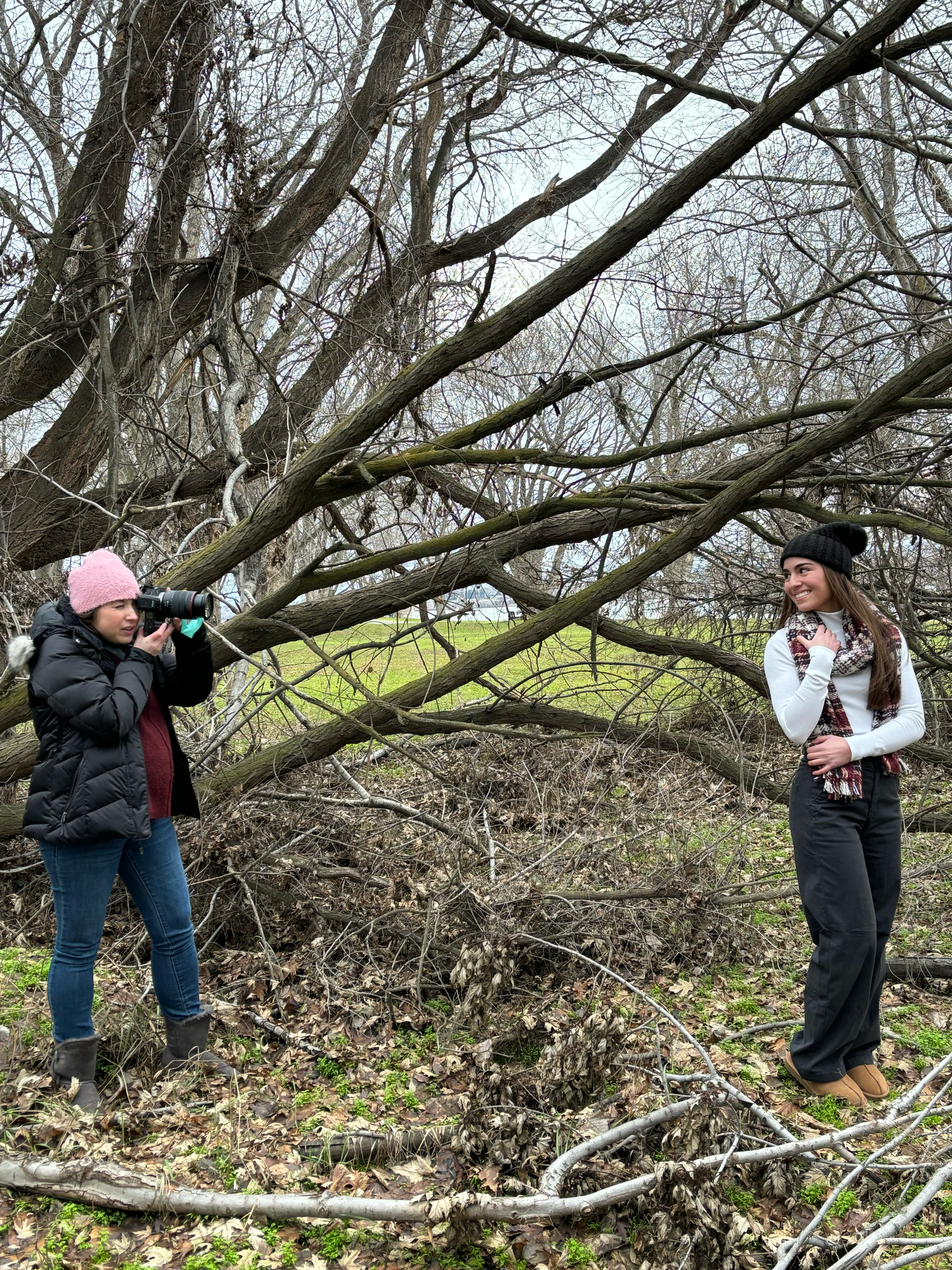 Two women in a bare winter forest, one photographing the other among leafless trees and fallen branches.