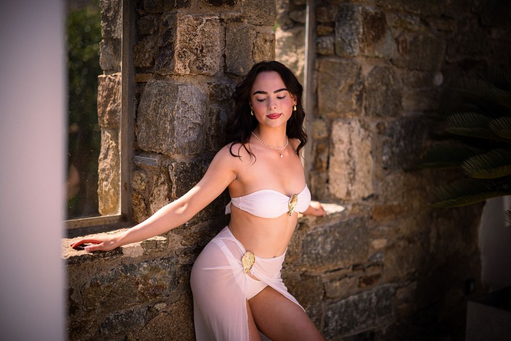 Woman in light pink swimwear poses elegantly against rustic stone wall backdrop.