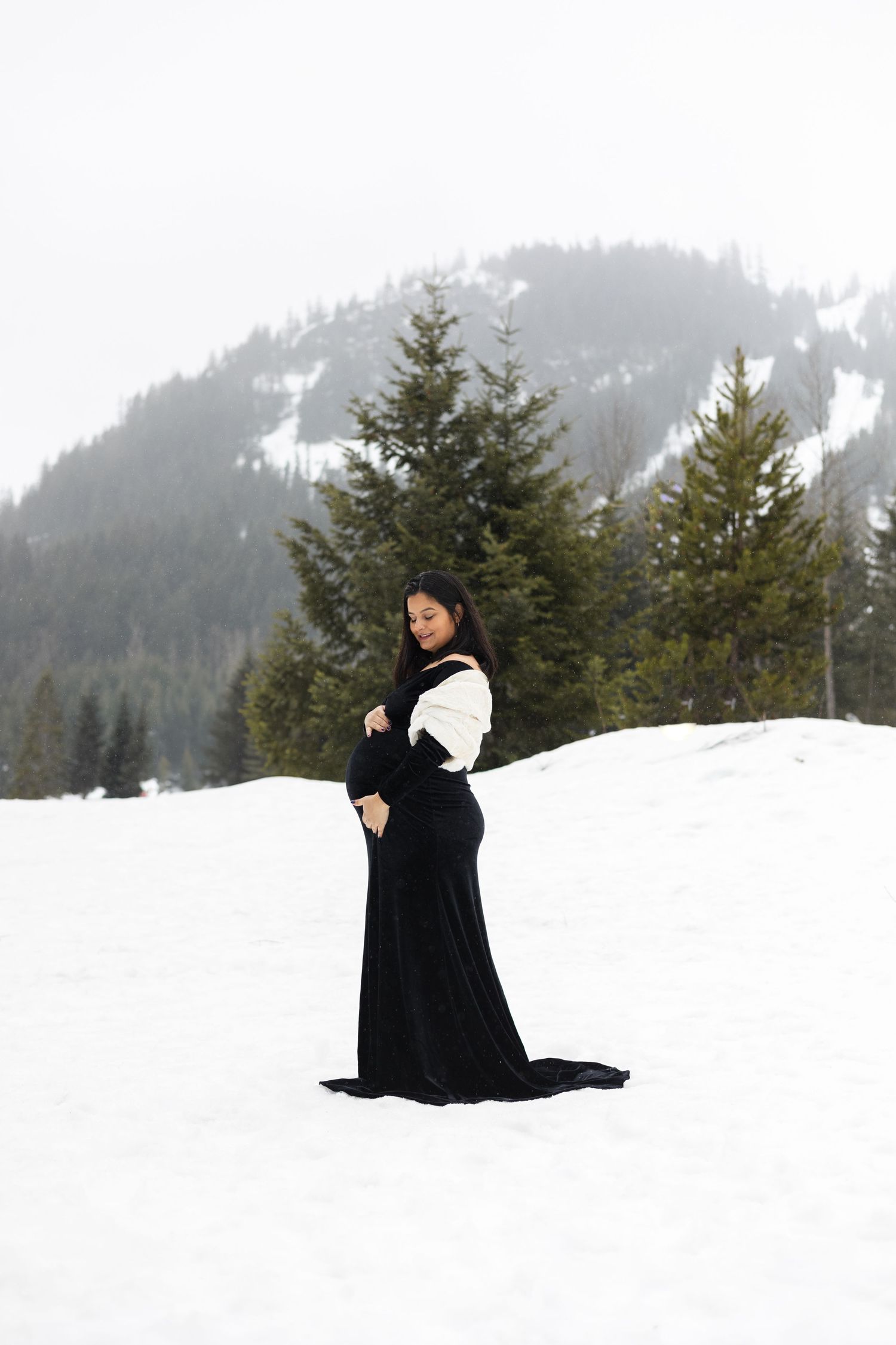 A person in a black winter coat poses dramatically in snowy mountains against evergreen trees and foggy peaks.