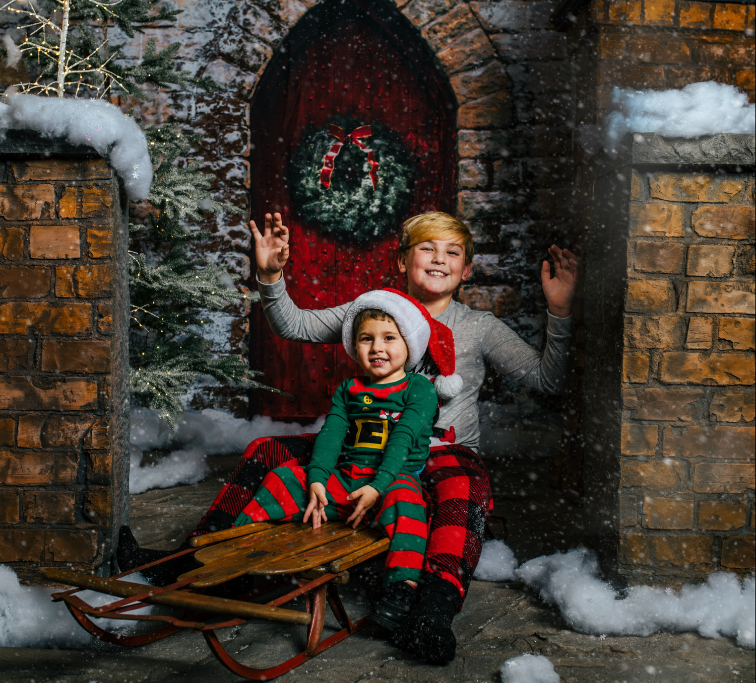 Two people in festive holiday outfits pose with a wooden sled against a snowy brick wall with a Christmas wreath.