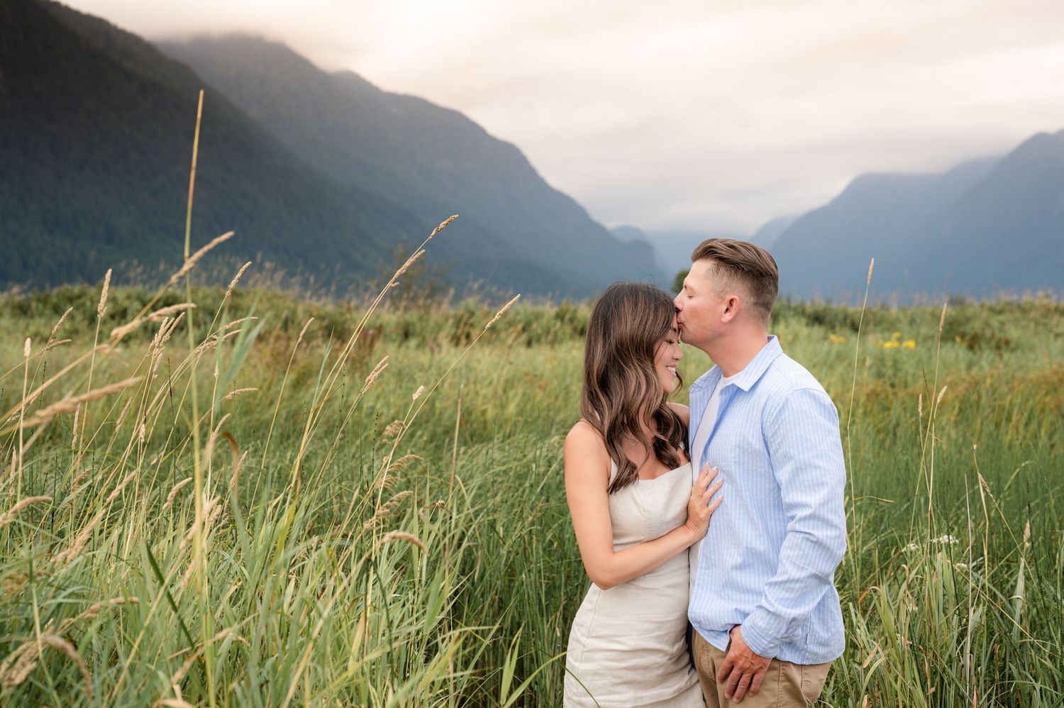 A couple embraces in a scenic mountain meadow with tall grass swaying in the wind during golden hour.