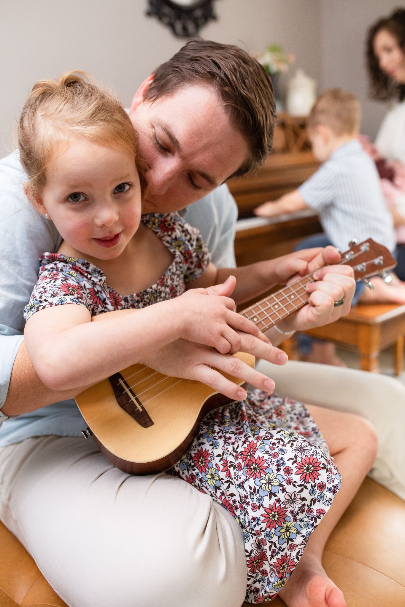 A child in a floral dress learns to play ukulele with guidance from dad sitting together on a couch.