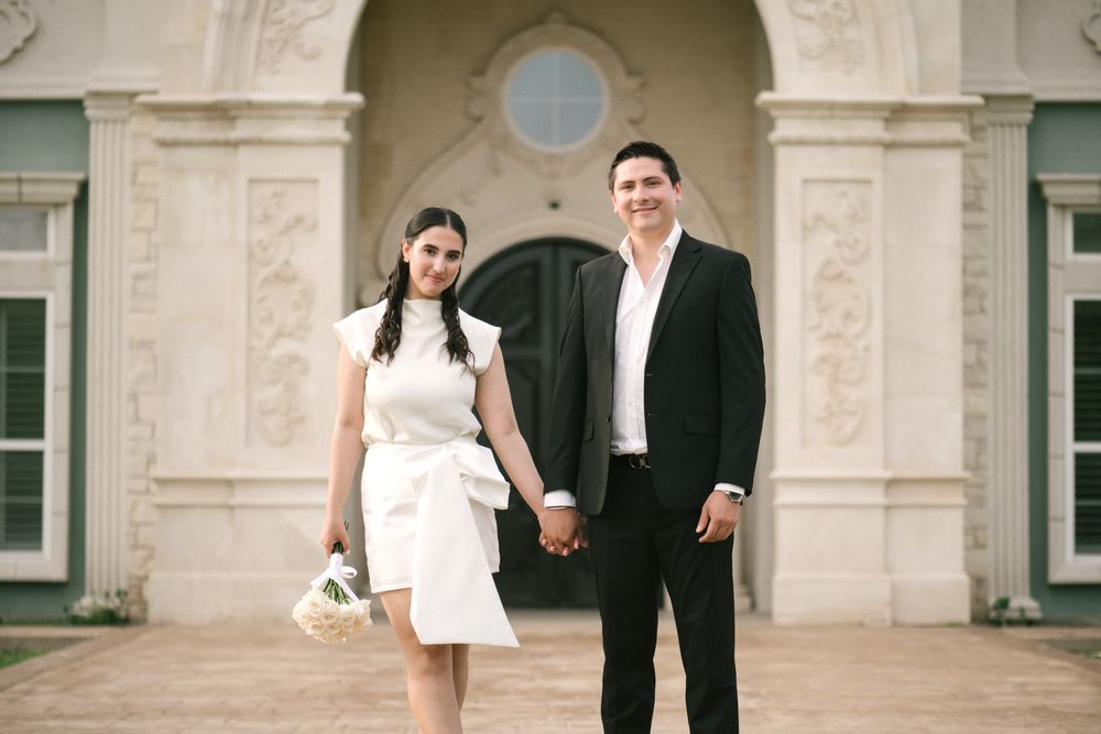 A wedding couple walks hand in hand along an elegant stone building exterior with arched windows.
