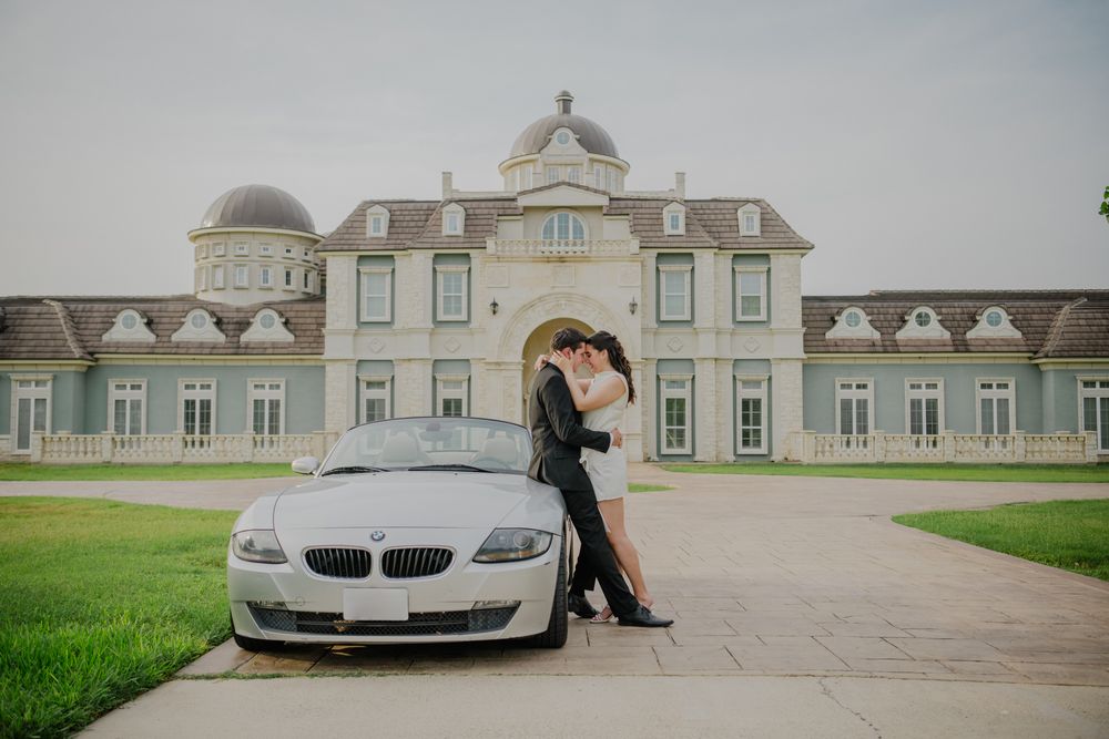 A couple embraces next to a silver BMW Z4 parked in front of an elegant European-style mansion with manicured grounds.