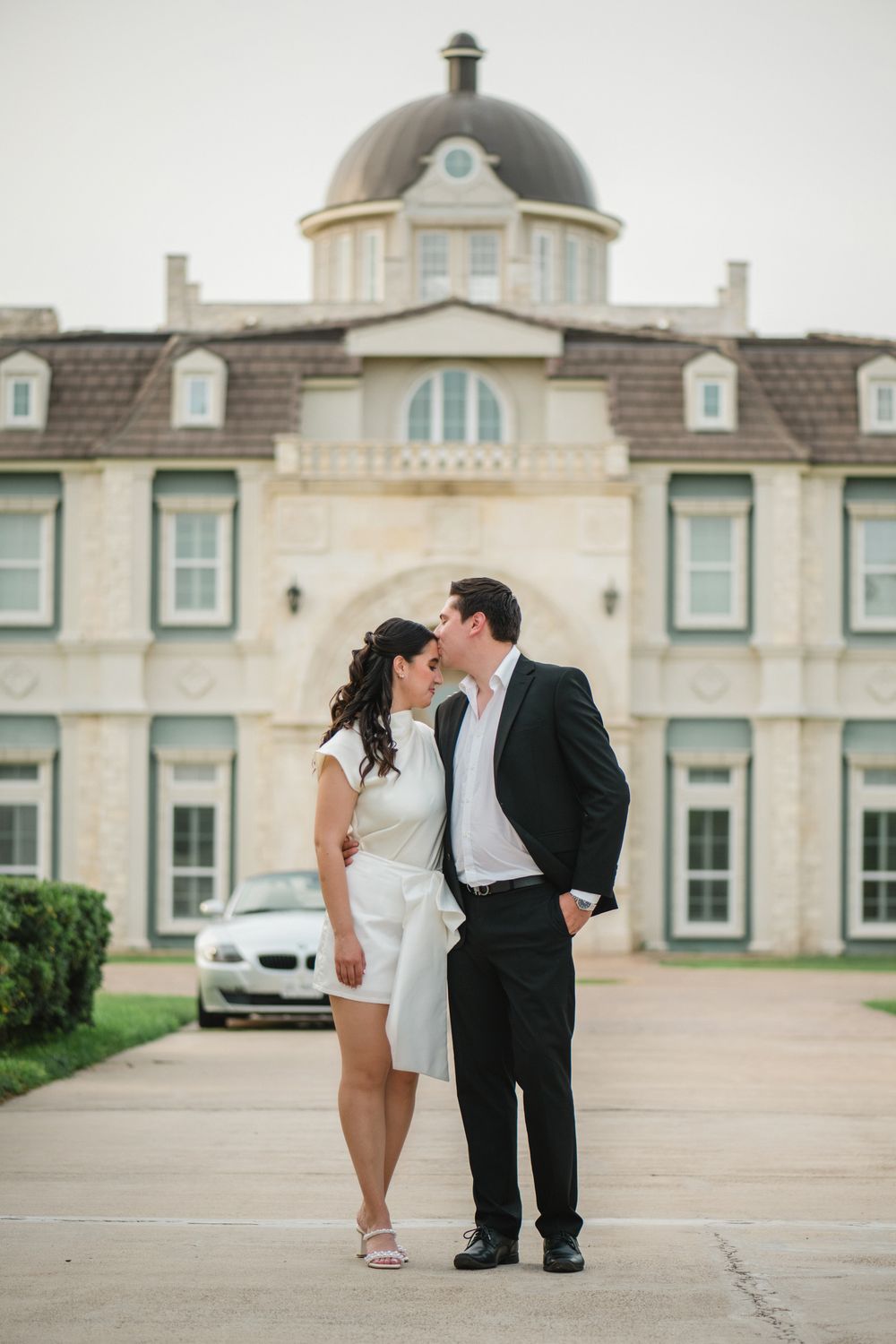 Stylish couple in formal attire poses together on stone courtyard of grand estate building.