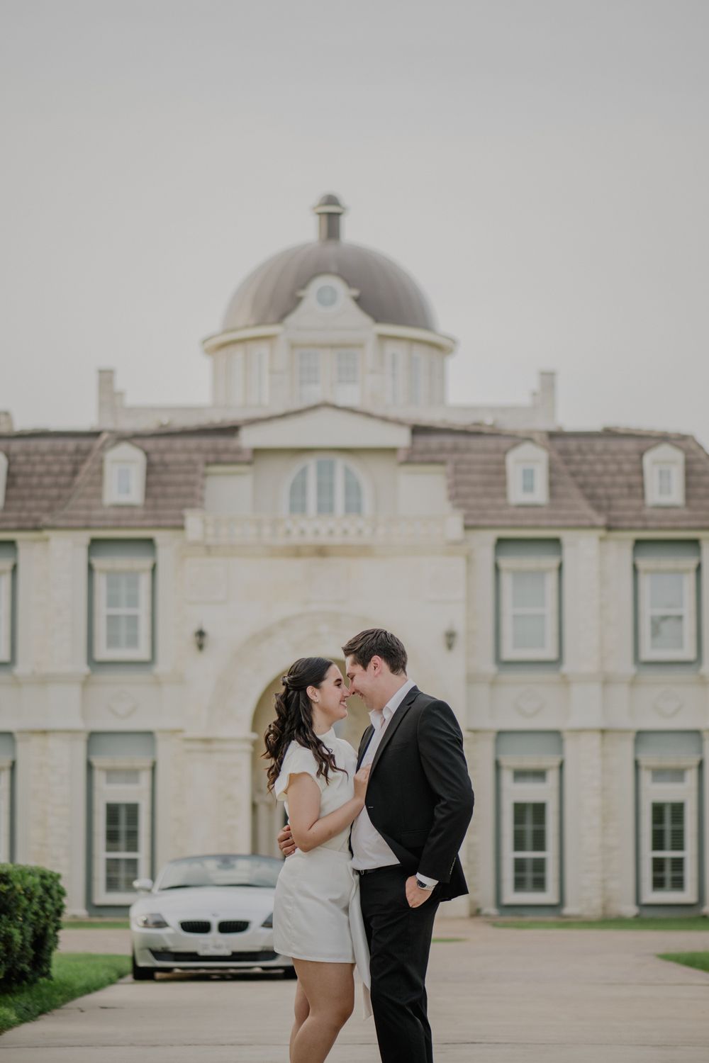 A romantic wedding couple shares an intimate moment in front of an elegant white mansion with domed architecture.