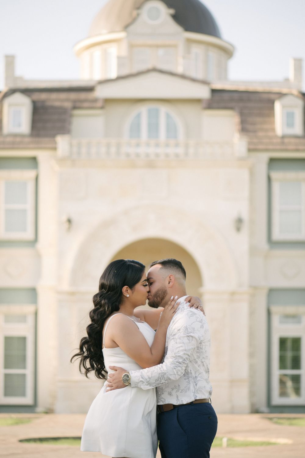 A romantic wedding couple shares an intimate moment in front of an elegant cream-colored mansion facade.