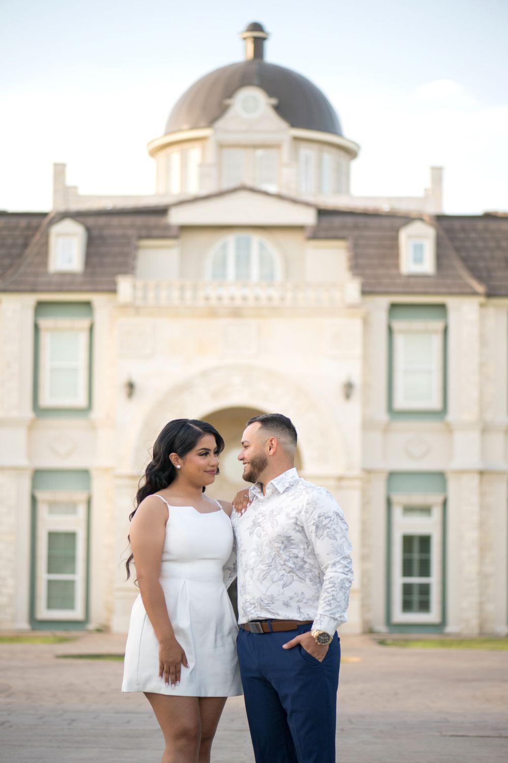 A romantic couple poses together in front of a grand stone mansion with domed architecture.