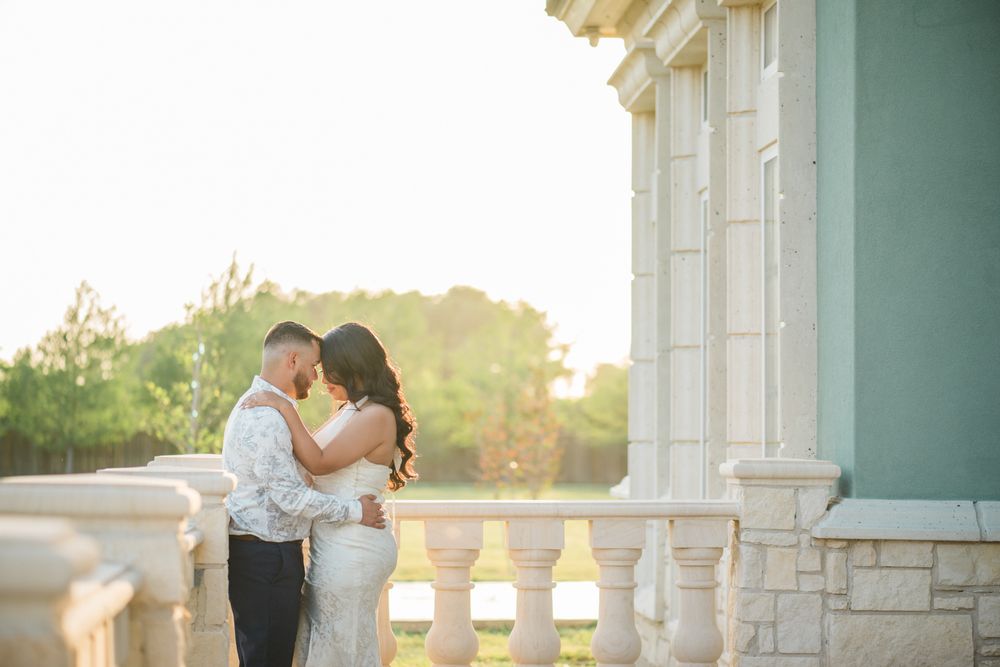 Romantic couple embraces on an elegant balcony with white columns during golden sunset hour.