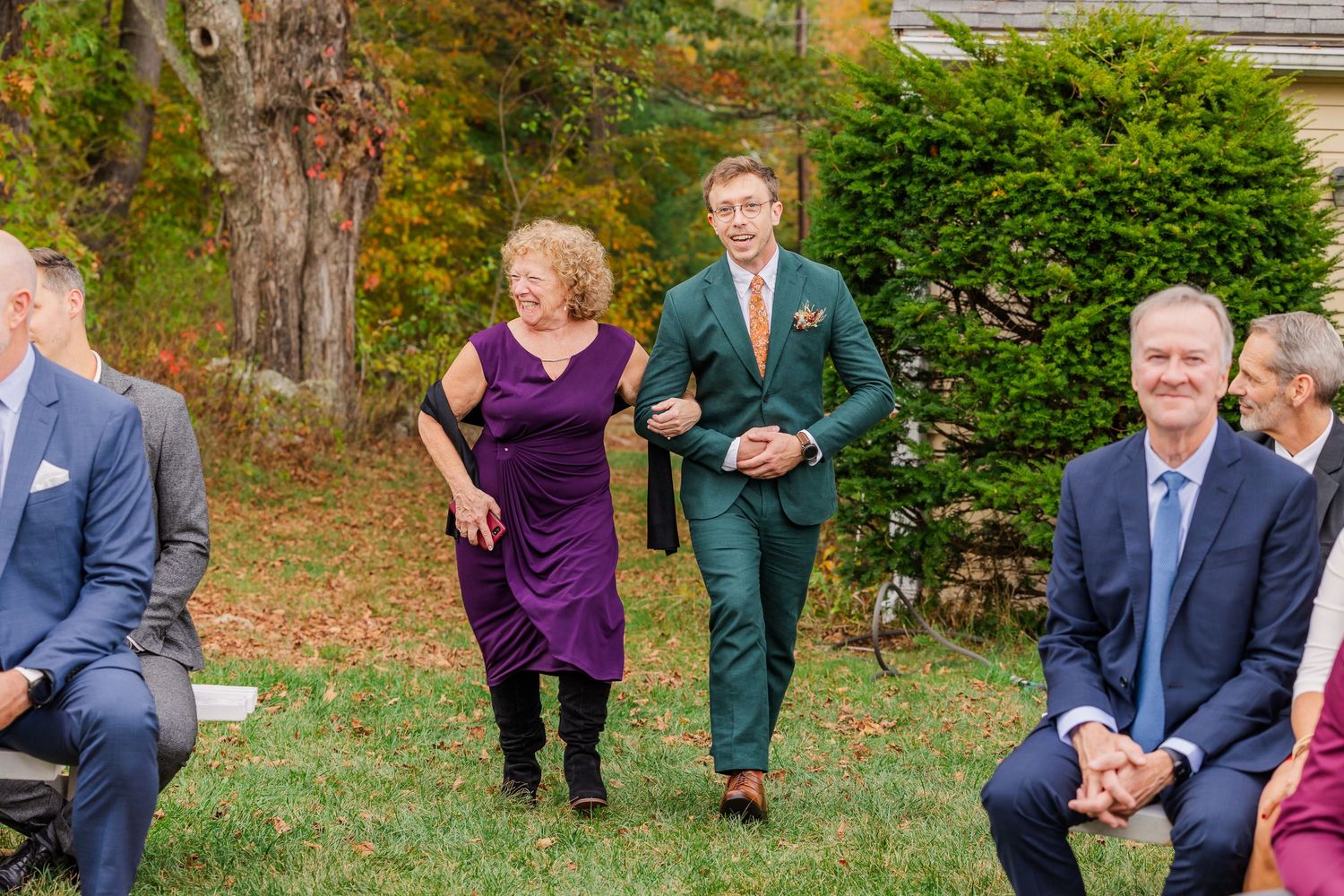 Wedding party shares candid laughter while posing outdoors on grassy lawn with trees in background.