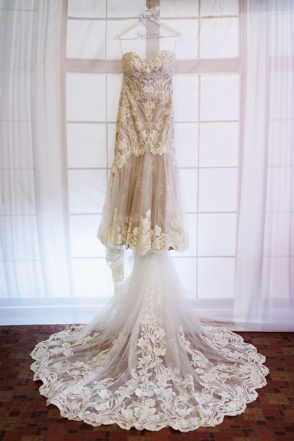 Elegant champagne lace wedding dress with sheer overlay and dramatic cathedral train displayed on wooden floor.
