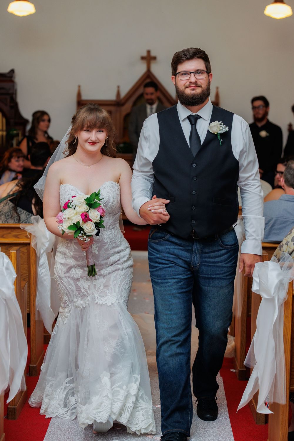 A wedding couple walks down a church aisle with white decorative bows adorning the pews.