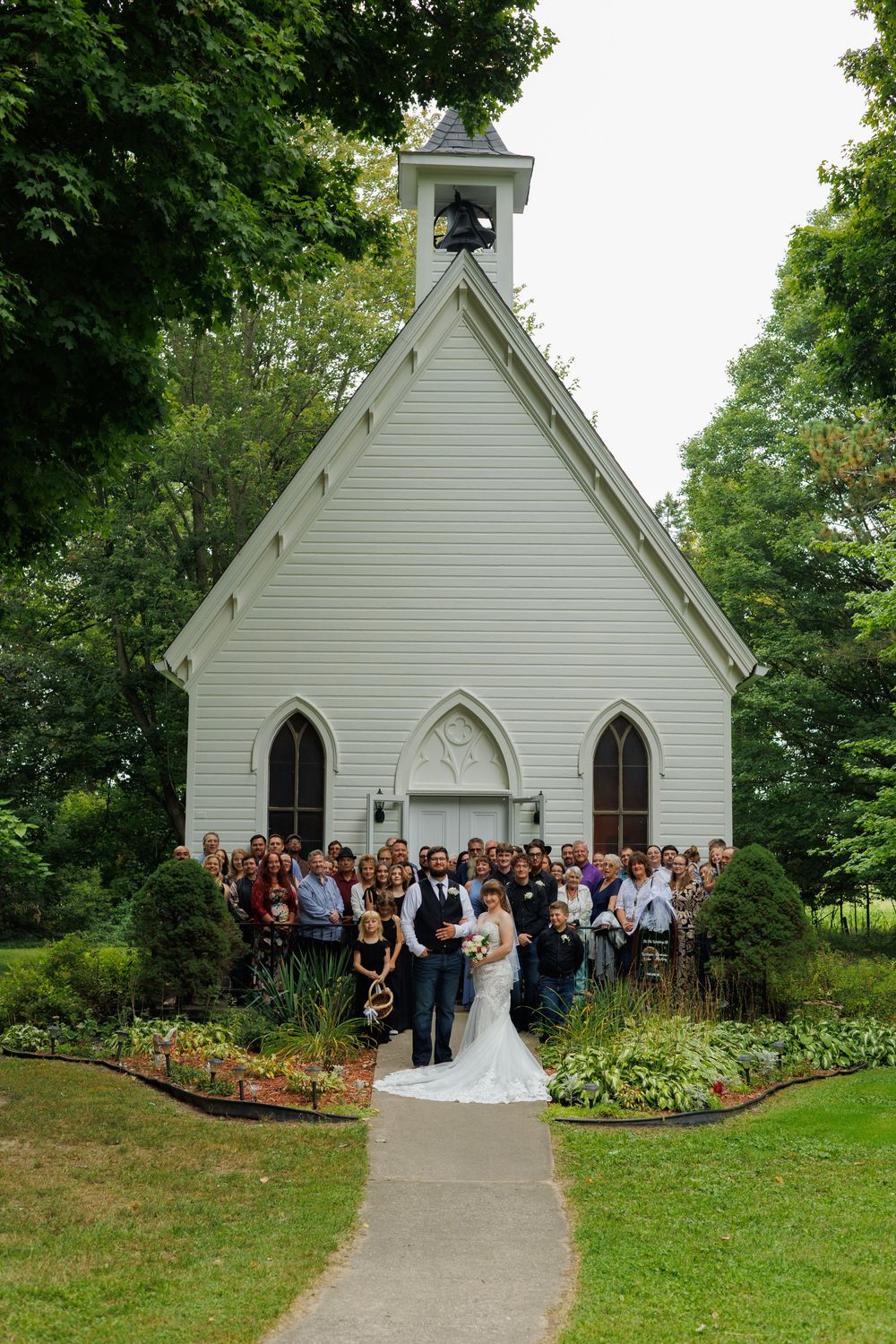 A white wooden chapel with Gothic details sits among lush greenery during an outdoor wedding ceremony.