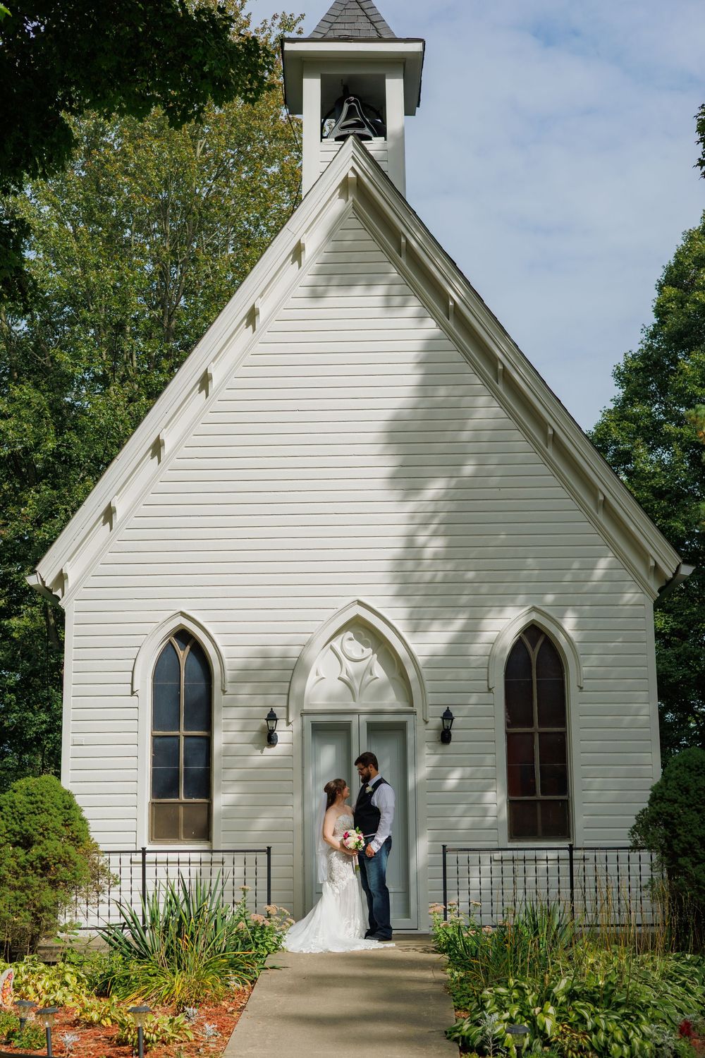 A charming white chapel with Gothic windows and a steeple stands against a blue sky surrounded by green trees.