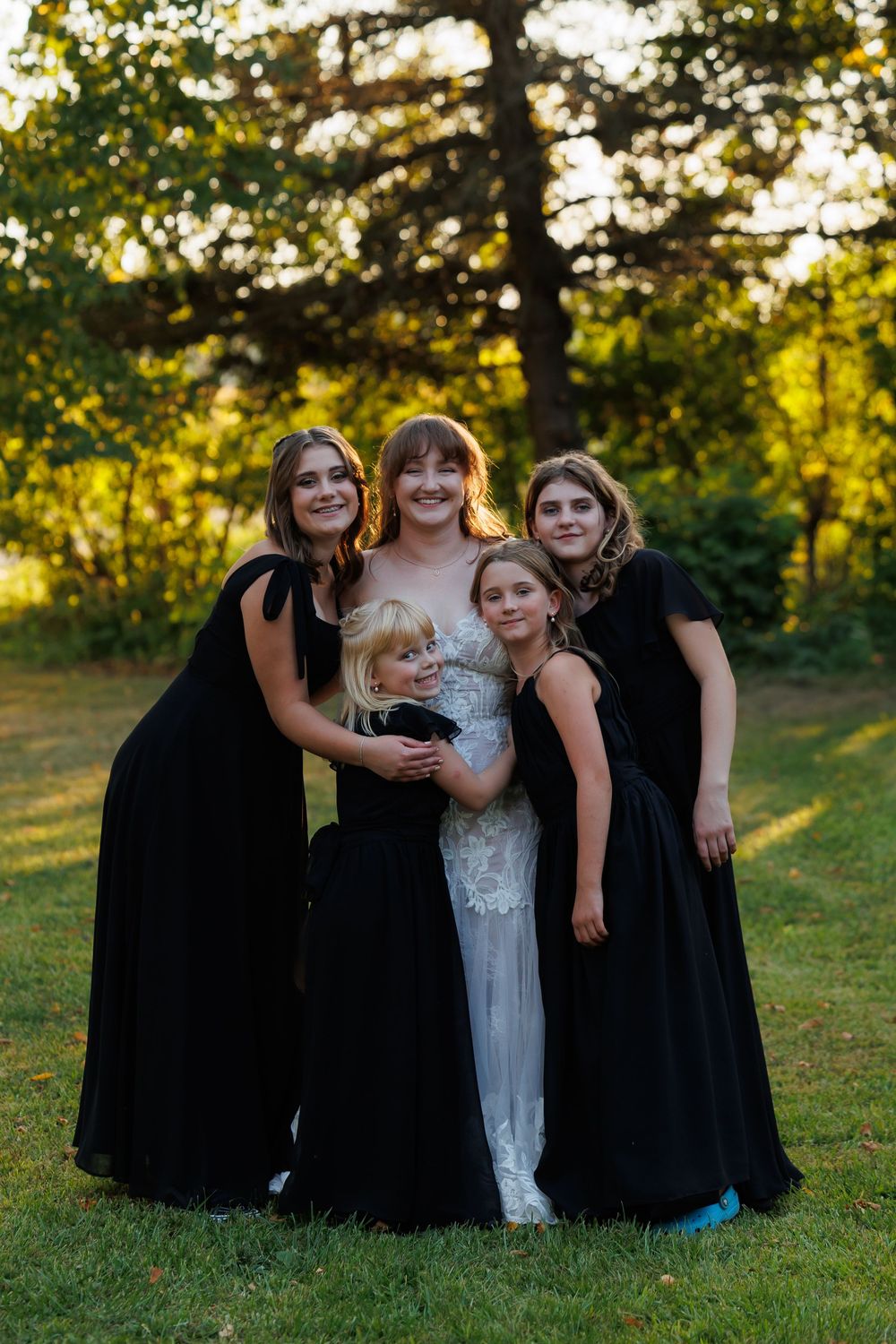 Wedding party poses on a grassy hillside during golden hour with bridesmaids in black dresses.