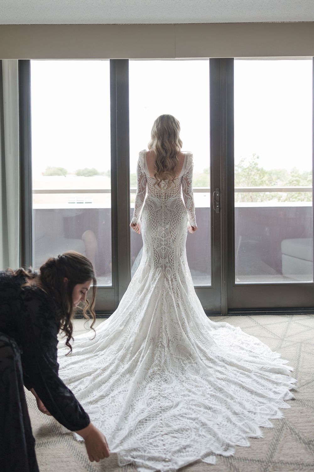 Back view of bride in lace wedding gown with long train standing at floor-to-ceiling windows while dress is adjusted.
