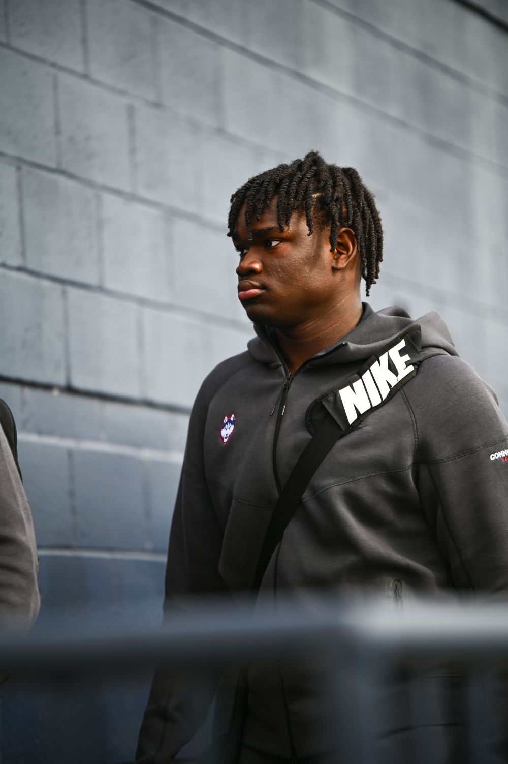 Athletic figure wearing Nike hoodie stands against concrete wall background.
