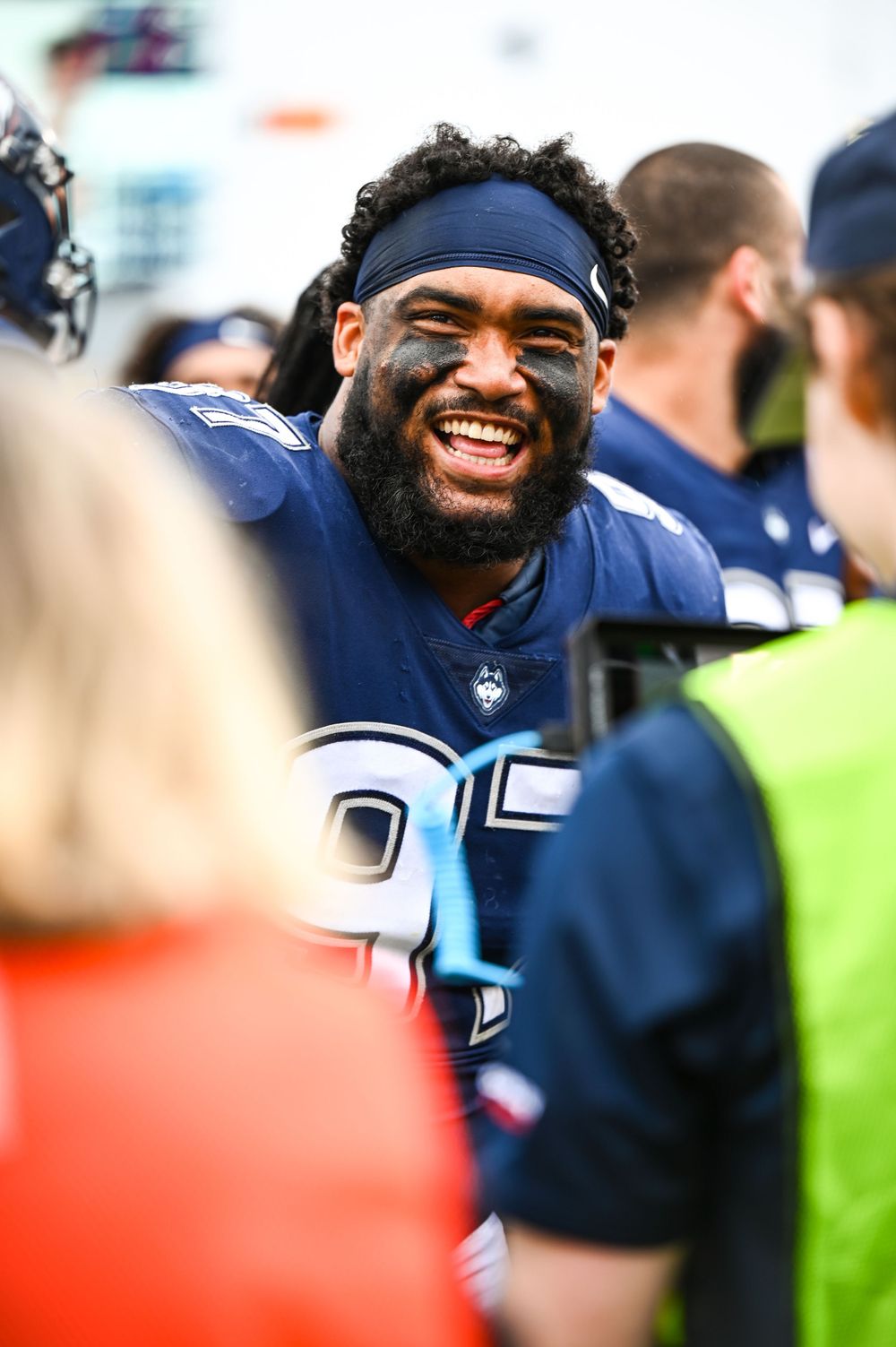 Smiling football player in UConn Huskies uniform with navy headband and face paint during a game.