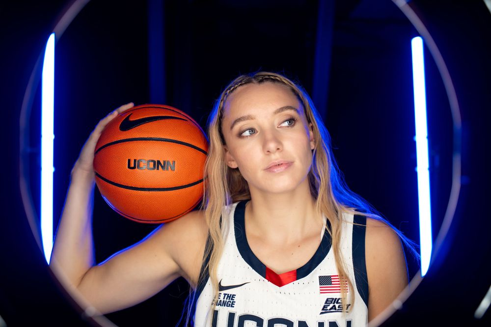 Basketball player, Paige Bueckers poses with ball against blue-lit background in UConn uniform.