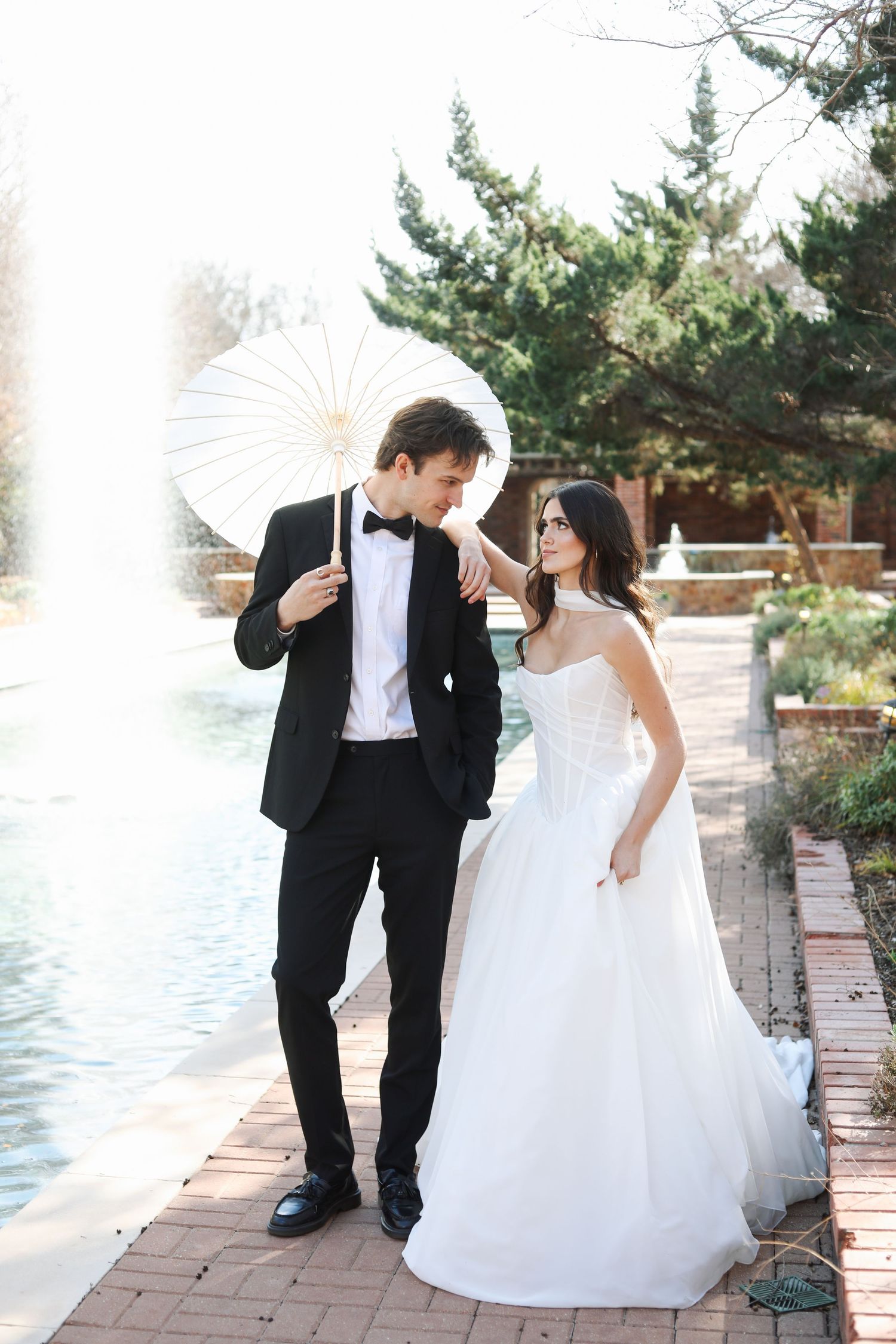 Romantic wedding photo session with white parasol on brick pathway by water.