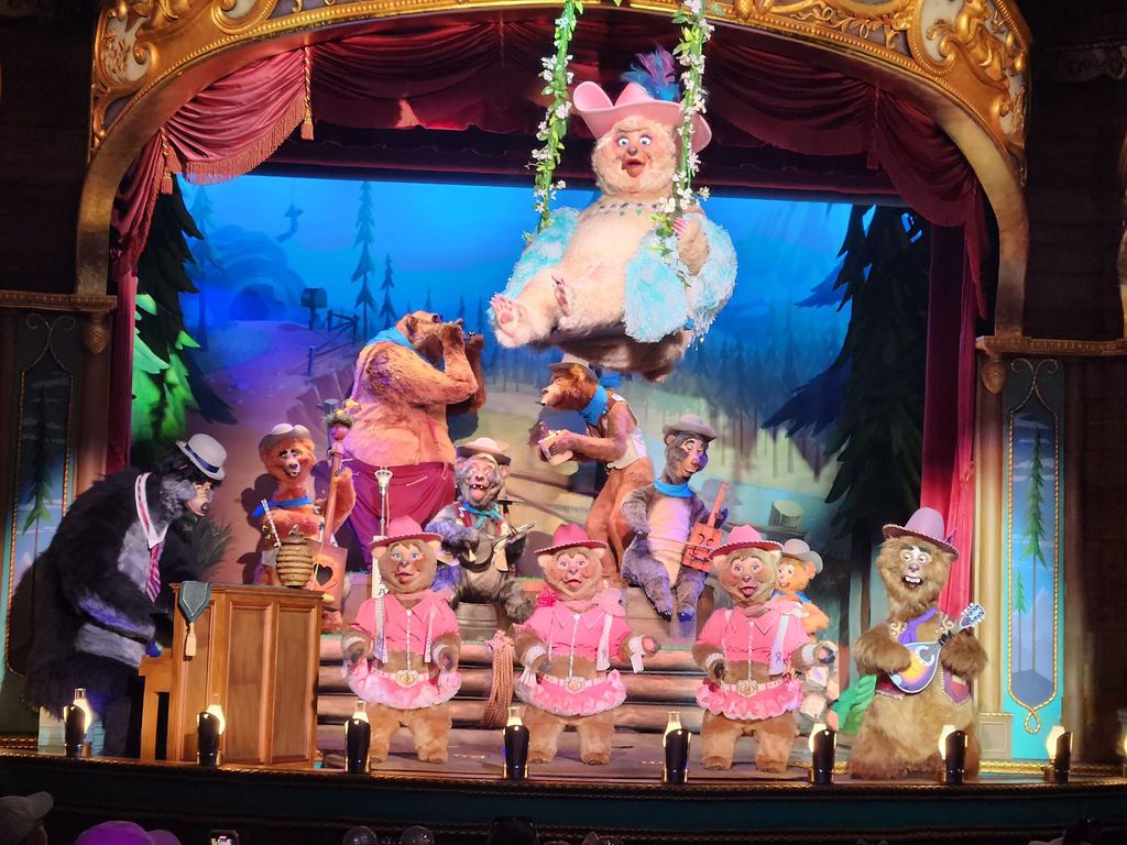 A colorful stage performance featuring puppet characters in pink outfits with elaborate theater curtains and lighting effects.