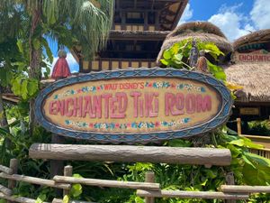 Vintage surfboard-style wooden sign with tropical theme and text reading Enchanted Tiki Room mounted on bamboo posts outdoors.